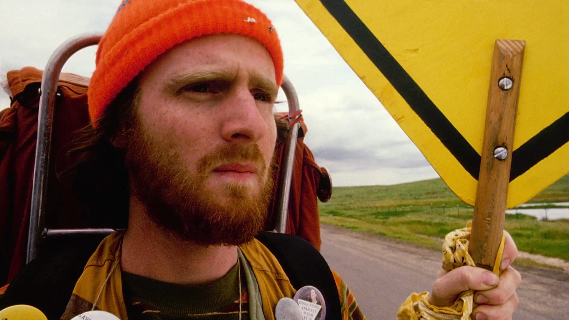A frustrated driver experiences a tense roadside encounter with a hitchhiker who signals them with a frog-themed warning sign. The interaction escalates into a frantic moment as the vehicle continues its journey across an open prairie landscape.