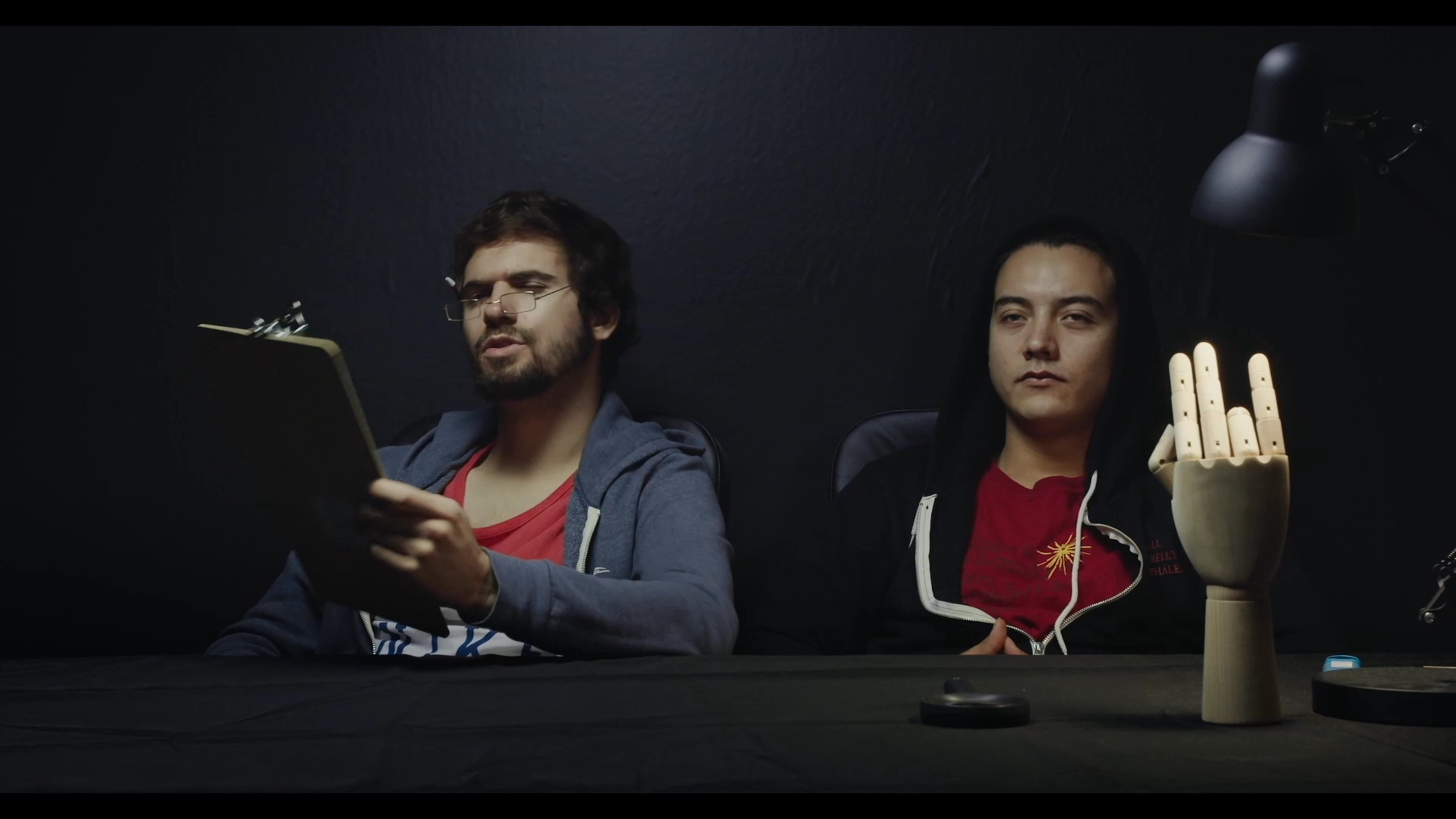 Two individuals engage in a lighthearted dialogue outside a study room before transitioning to a dark, stylized interview set featuring a wooden mannequin hand.