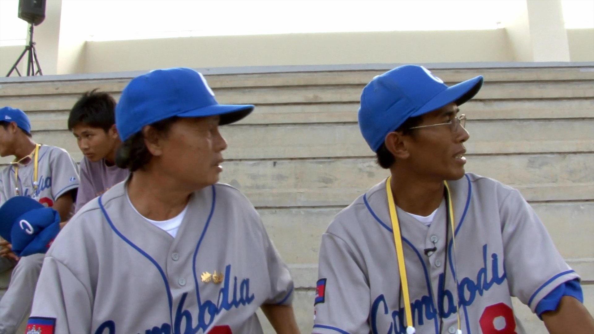 Two men conduct an interview in an indoor lounge setting, followed by footage of baseball coaches in Cambodia discussing tactics and logistics on the bleachers of a stadium.