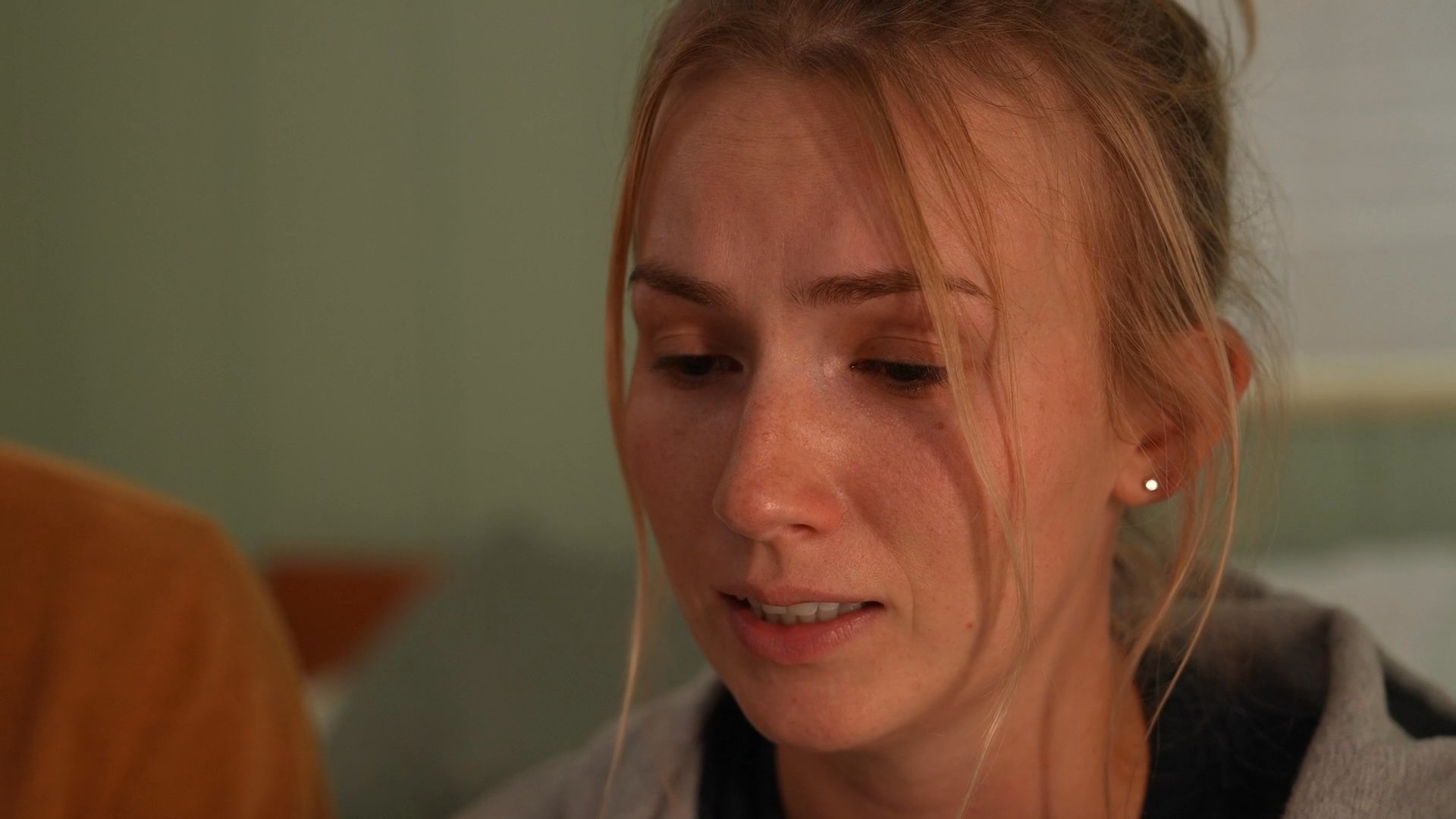 A couple sits together on a bed in a somber, intimate exchange as the woman experiences visible emotional distress. The scene shifts to her lying down while her partner remains nearby, emphasizing a heavy, shared moment of vulnerability.