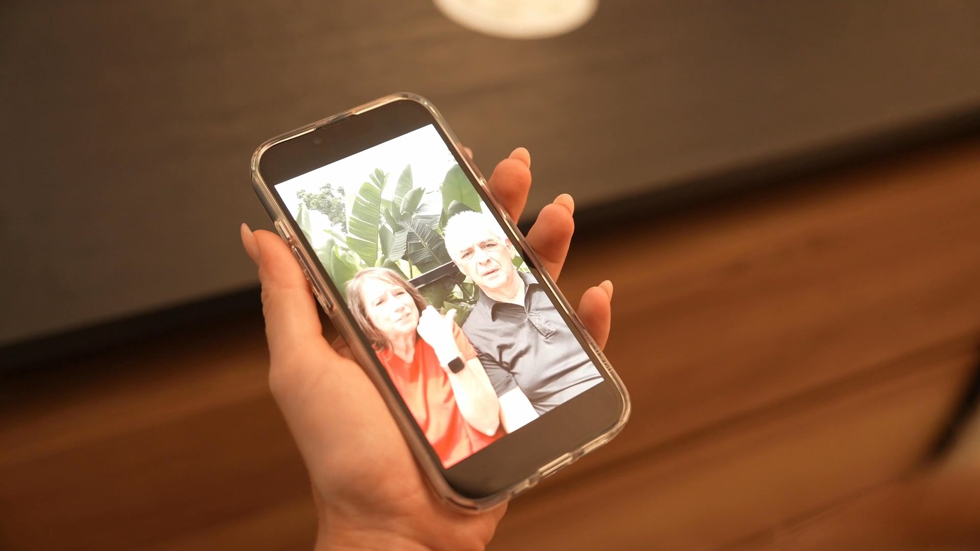 A person holds a smartphone displaying a photograph of a couple while standing in a room. The individual navigates the mobile interface while observing the image content.