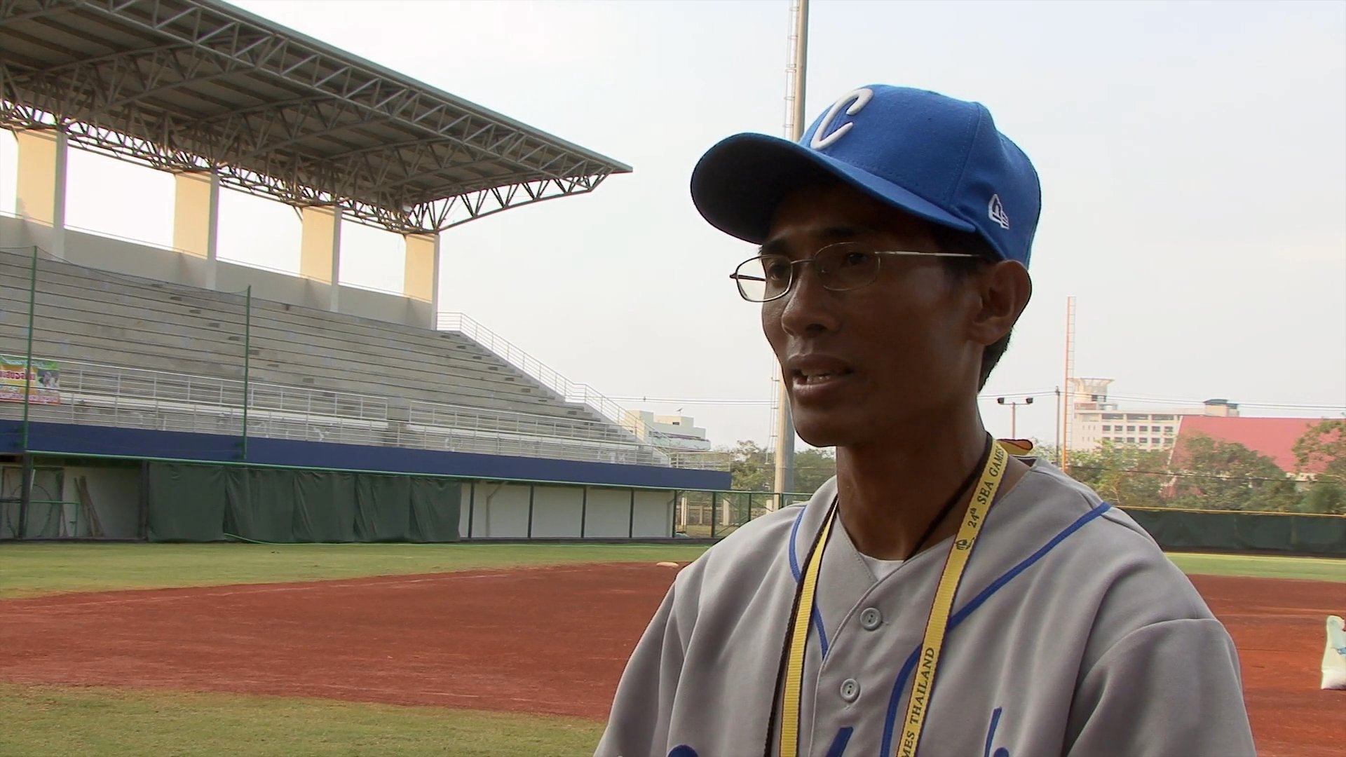 A coach wearing a baseball uniform speaks during an interview at an empty outdoor stadium.