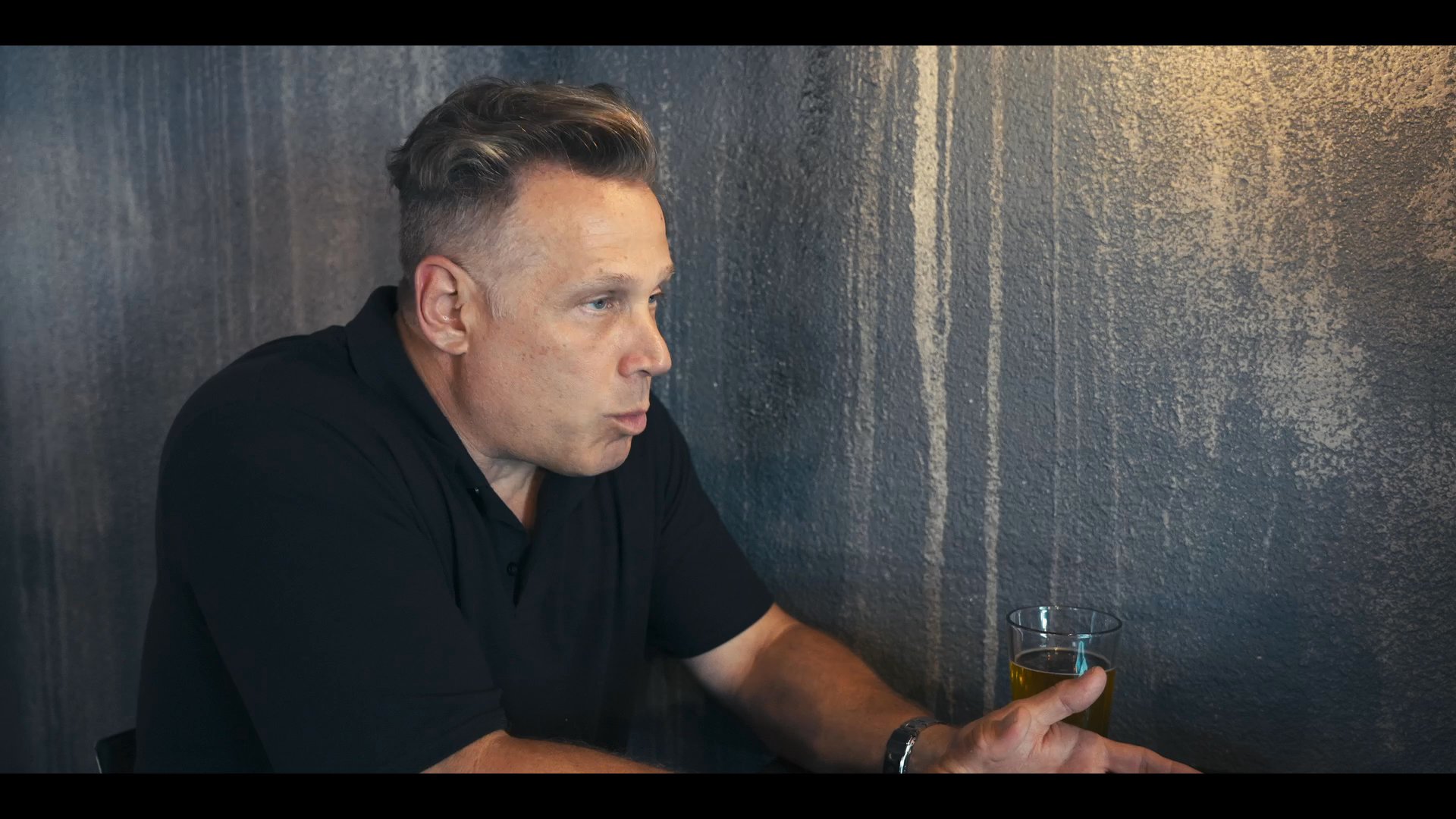 Two men engage in a focused, professional dialogue while seated against a textured wall. They alternate between speaking and listening, maintaining an attentive and composed demeanor throughout the exchange.