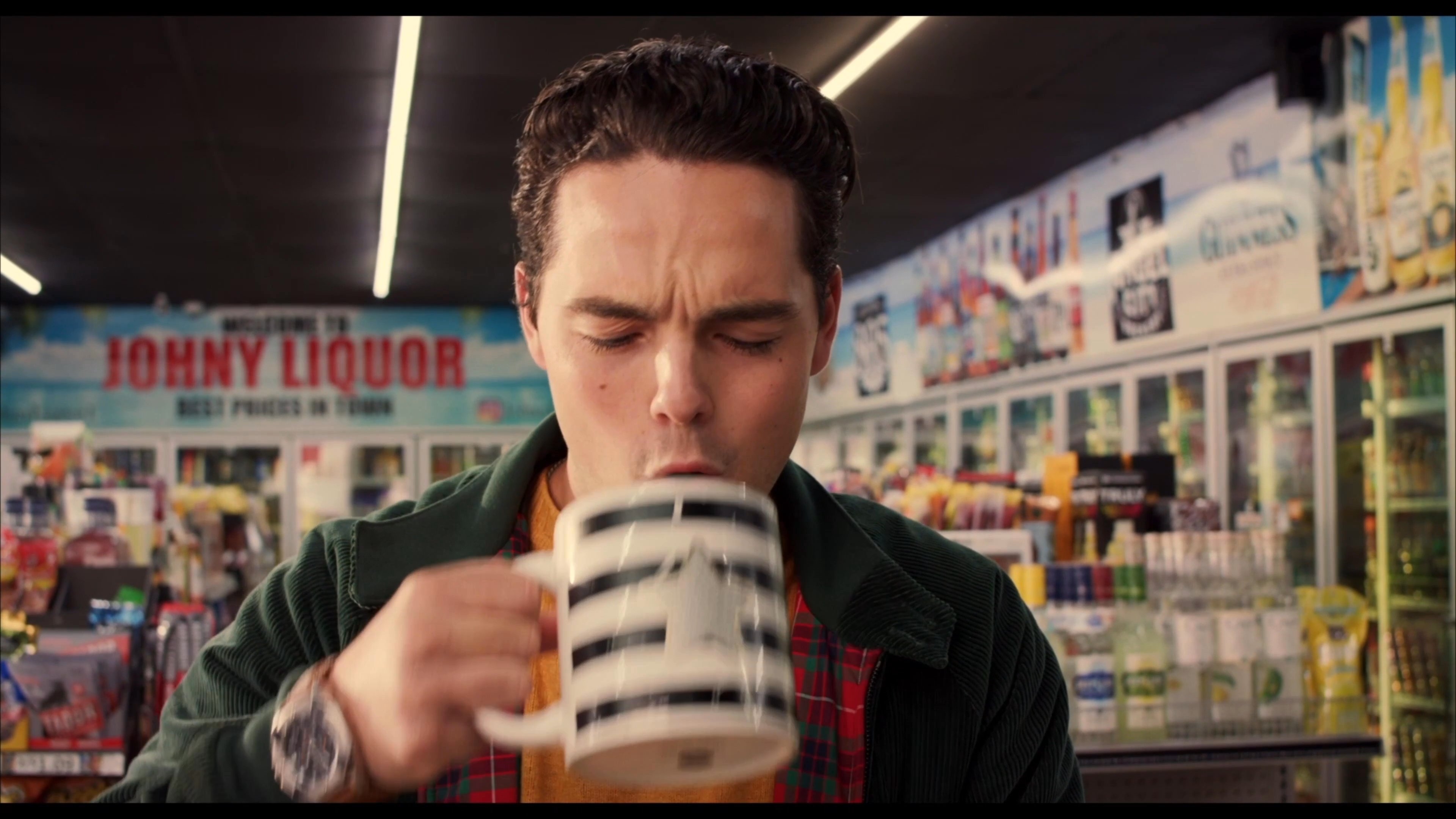 A man samples a beverage from a novelty mug inside a liquor store while a store clerk looks on. The interaction highlights a lighthearted, casual atmosphere in a retail setting.