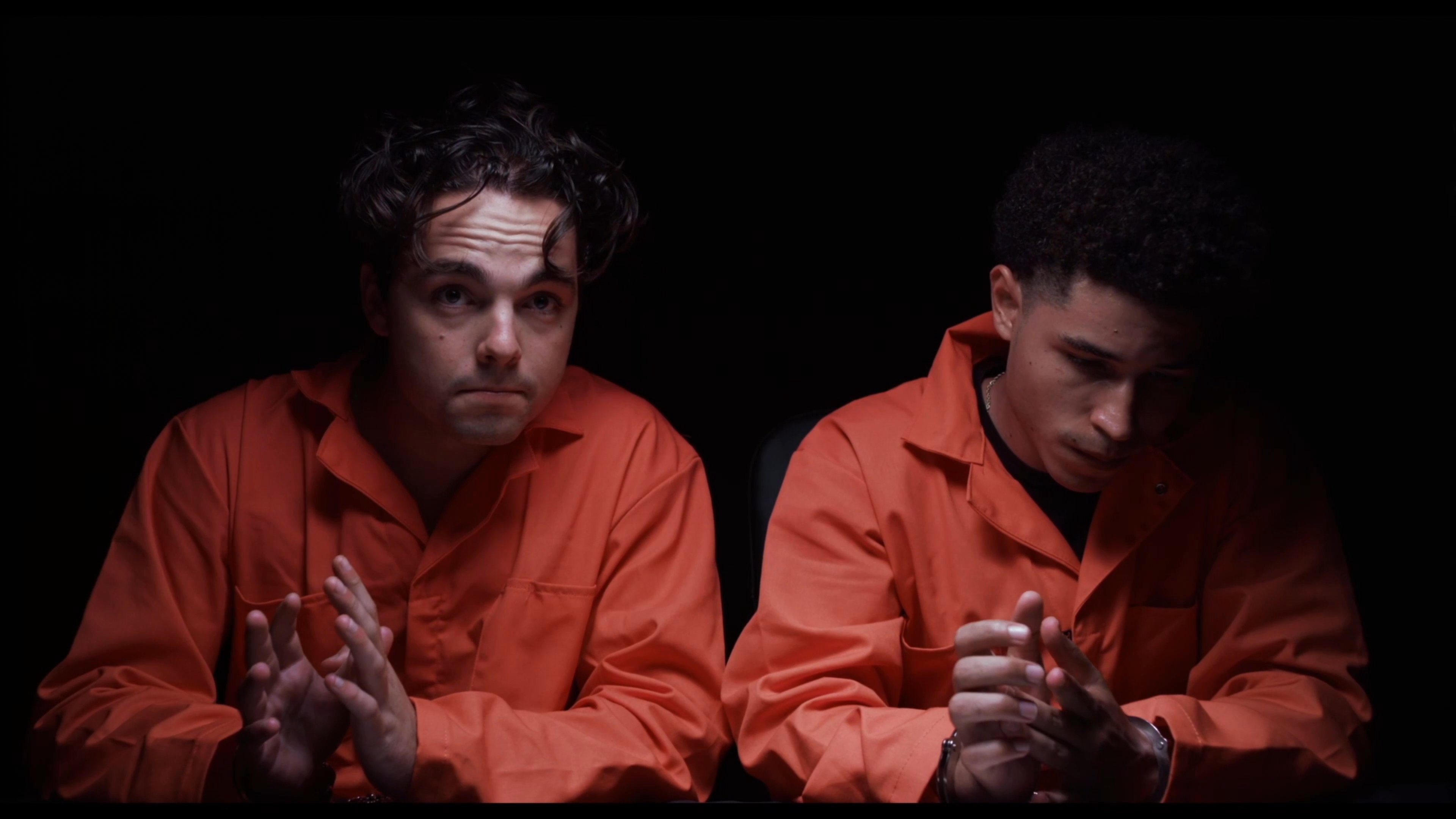 Two detained individuals in orange jumpsuits sit in a dark room under observation by a federal agent. The suspects appear anxious while interacting with their handcuffs during the high-tension encounter.