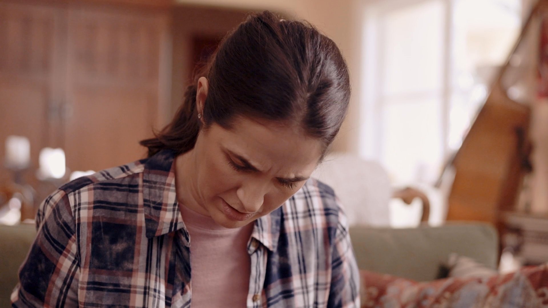 A woman sits in a living room, appearing visibly distressed and overwhelmed while a man stands nearby, attempting to engage her in a conversation.