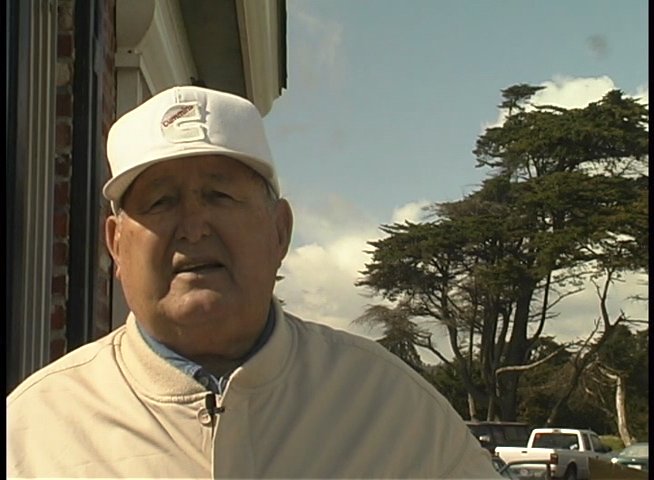 An older man wearing a baseball cap speaks directly to the camera in an outdoor setting.