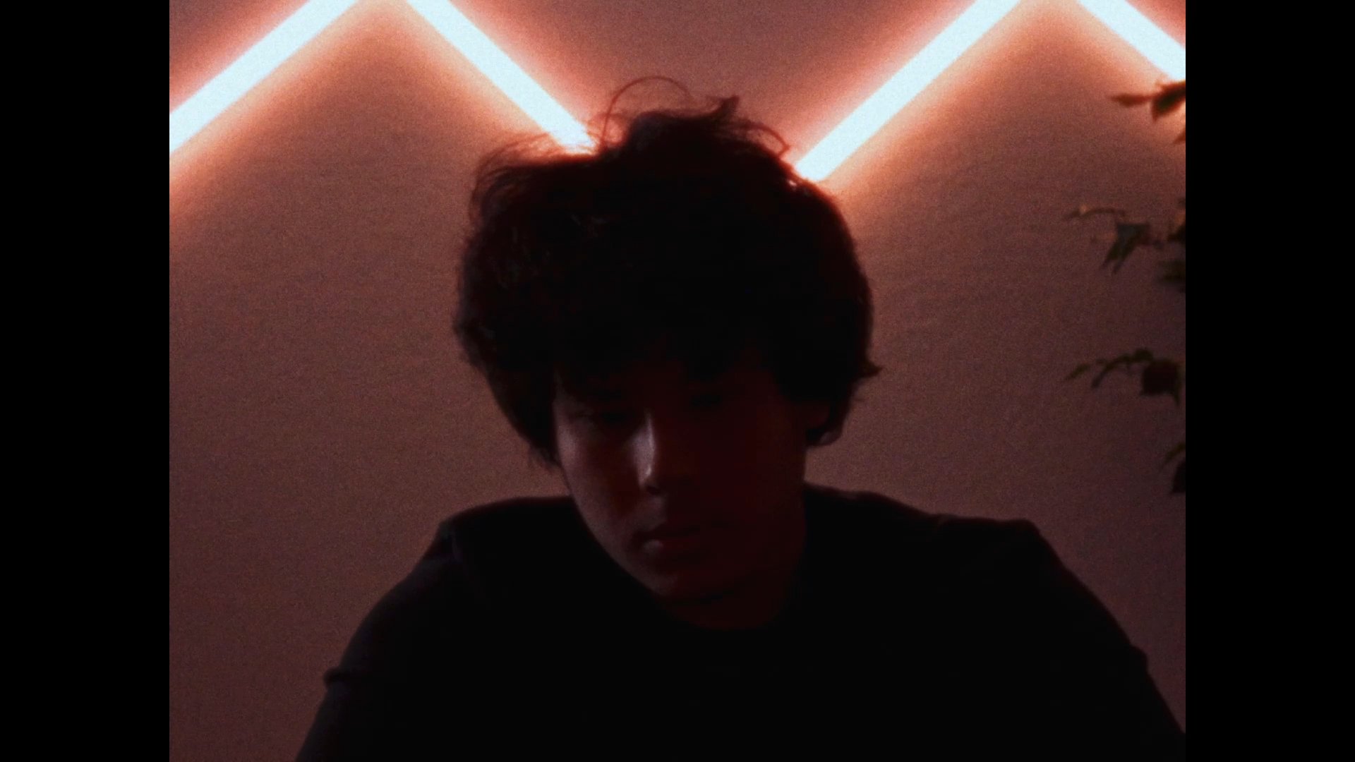 A young man sits contemplatively on his bed in a dimly lit room accented by neon lighting. He transitions from a seated position to lying down, suggesting a moment of reflection or rest.