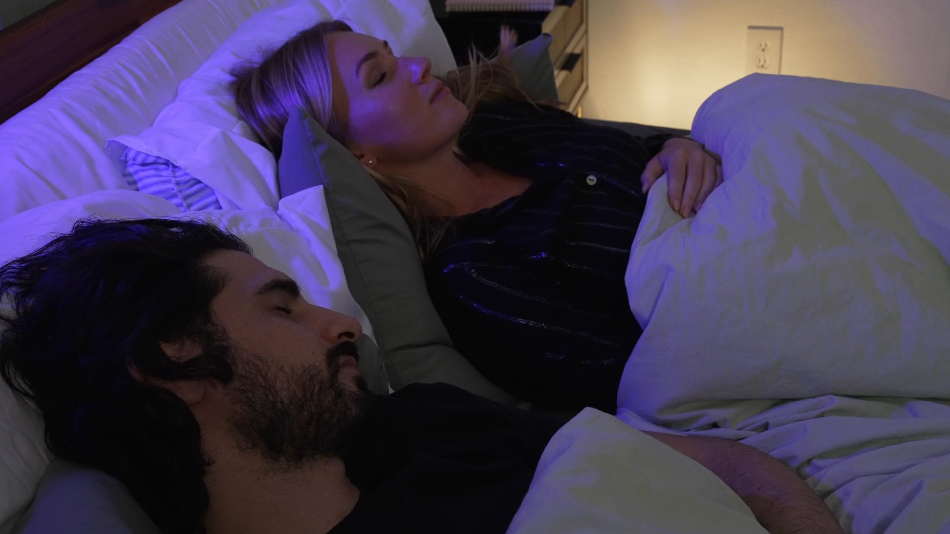 A man and woman engage in a quiet conversation while lying in bed together before the scene transitions to a memory or different location. The intimate exchange highlights a moment of personal connection in a domestic setting.