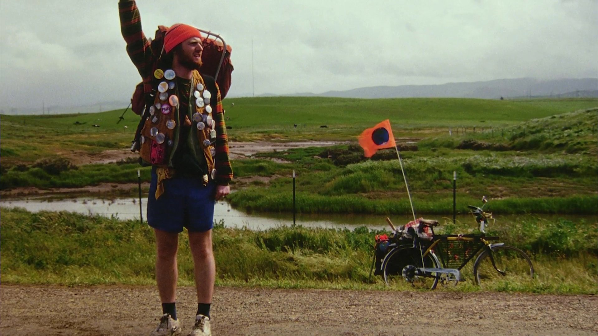 A backpacker standing on a rural roadside attempts to secure a ride by signaling to passing vehicles. The scene captures the solitary and expectant nature of long-distance hitchhiking through an open landscape.