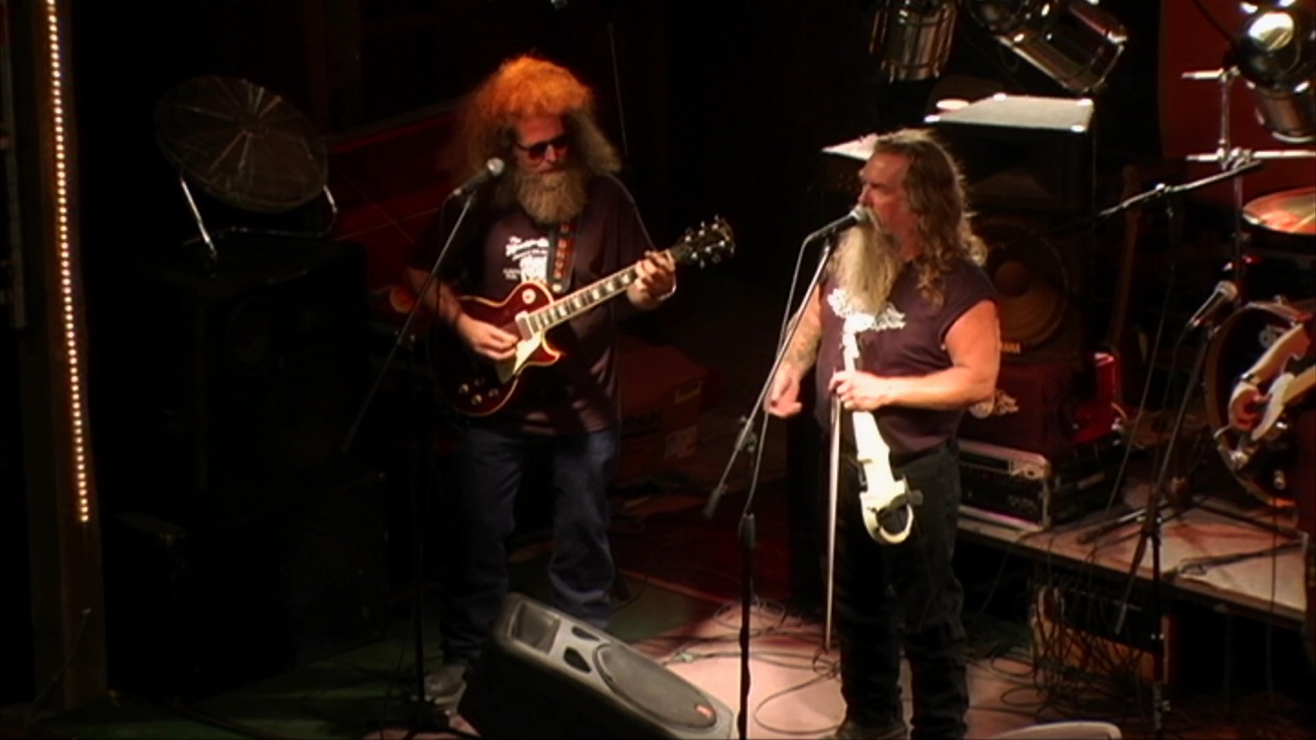 A long-haired, bearded singer performs live on stage, accompanied by a guitarist. The duo delivers an intimate musical set in a dimly lit, professional performance space.