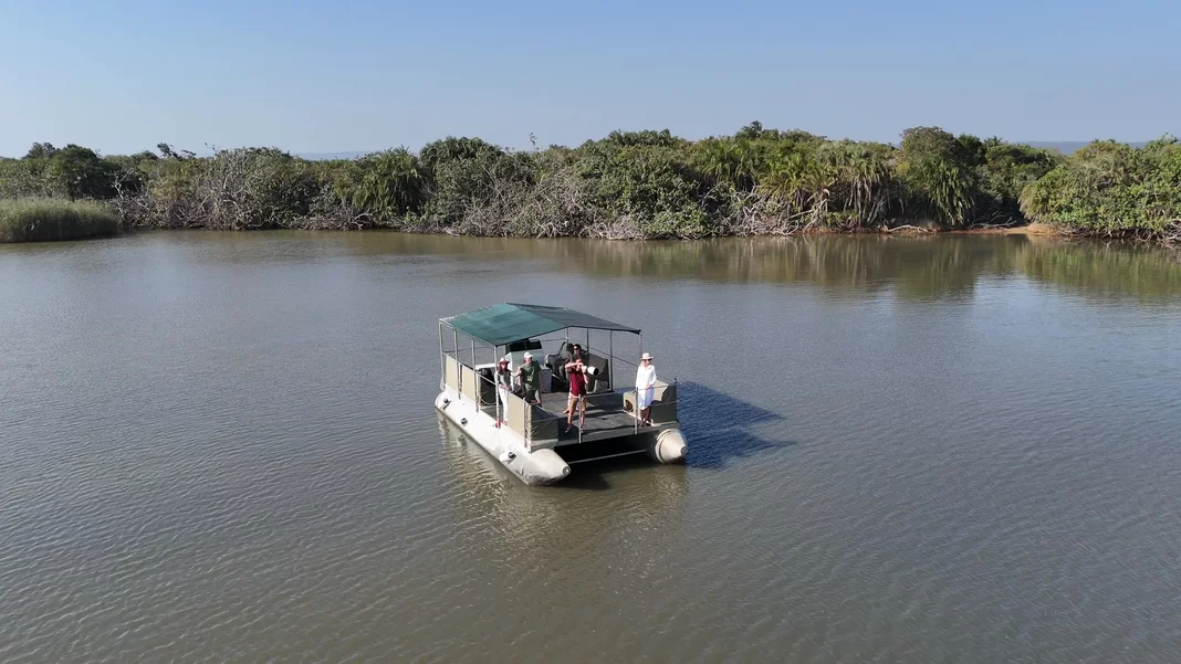 Guests line the rail of the canopied pontoon cruise boat on the flat water of the narrows, one photographing the green shoreline with a long lens.