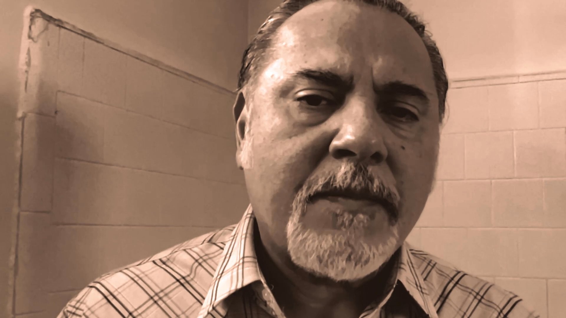 A middle-aged man with a graying beard delivers a somber, direct address to the camera in a sepia-toned environment. His steady gaze and measured movements suggest a serious or reflective narrative context.
