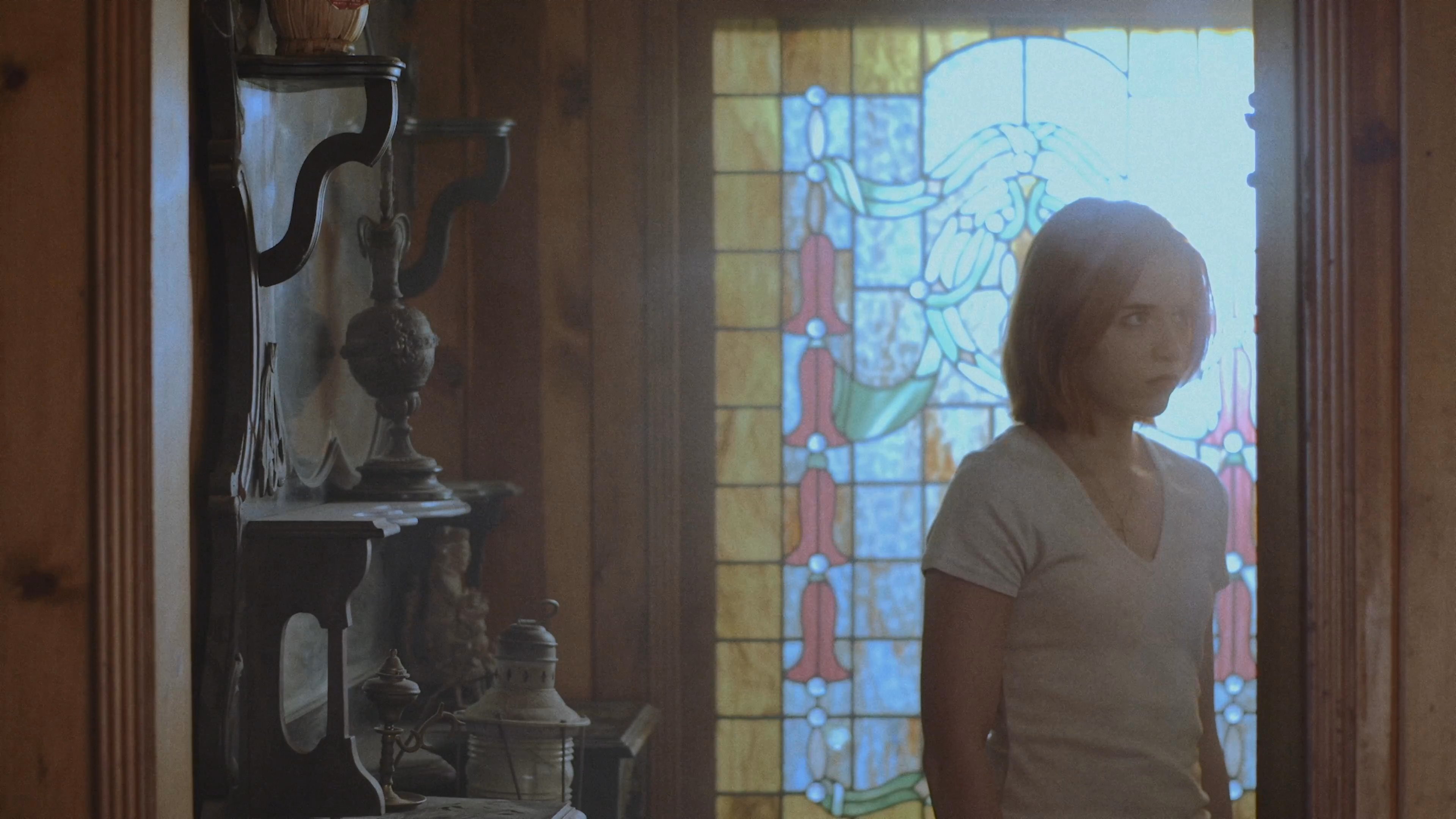 A woman stands before a stained glass window before walking across a dimly lit, rustic living room. The scene emphasizes a quiet, reflective atmosphere within a residential setting.