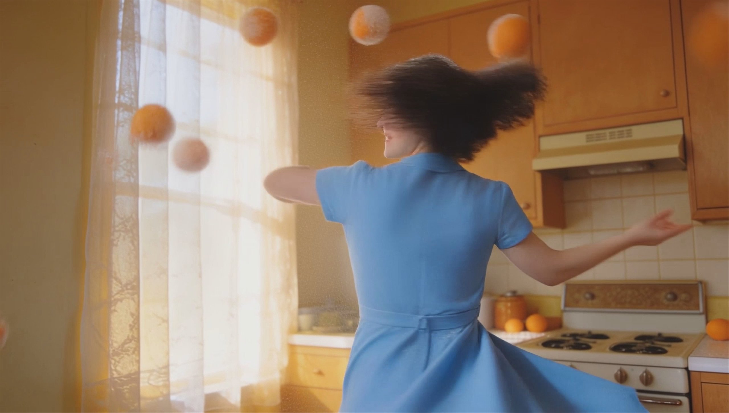 A woman in a vintage blue dress juggles sugar-dusted oranges in a sunlit retro kitchen.