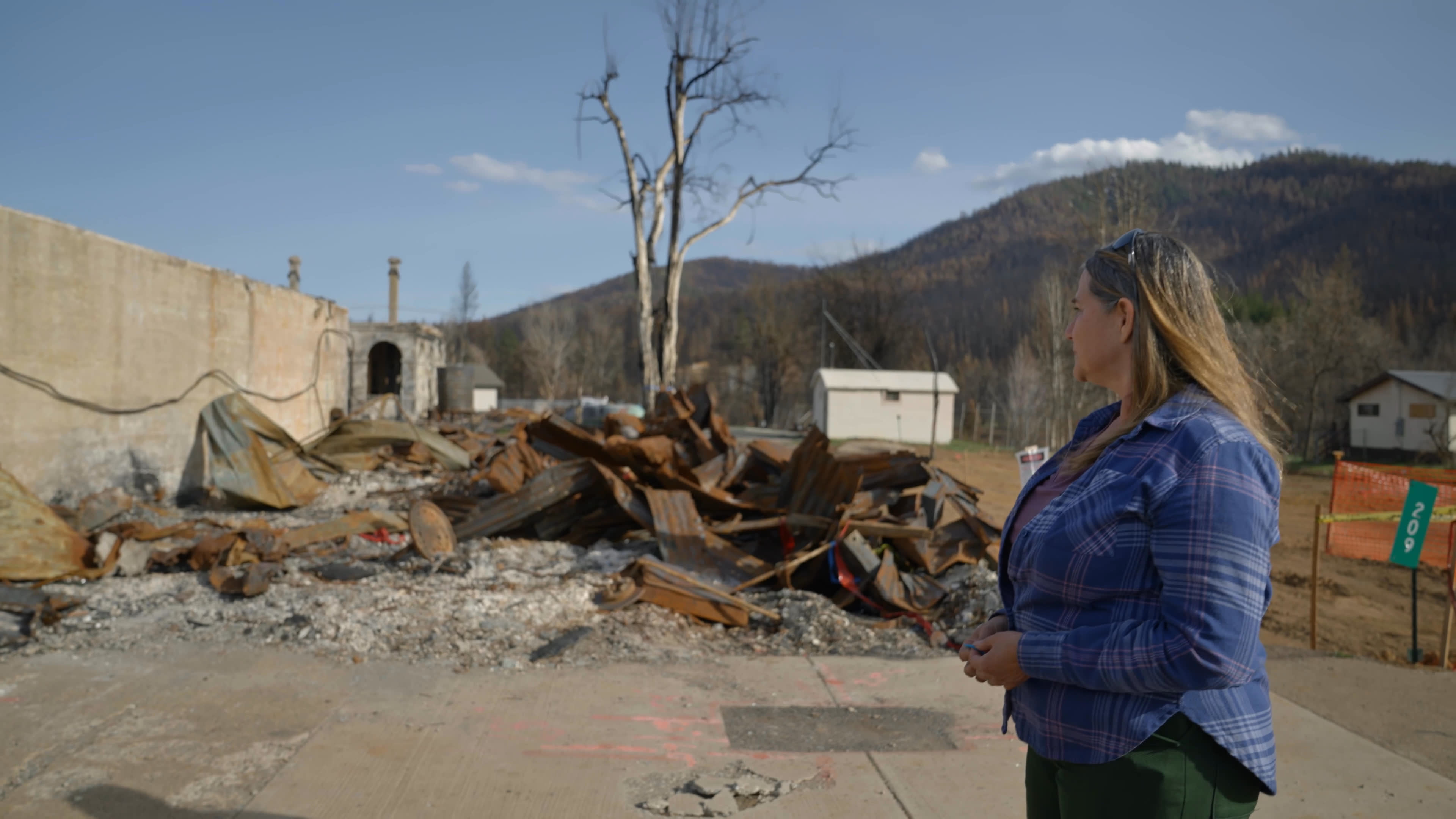A woman revisits fire-ravaged town ruins, with charred debris and damaged buildings surrounding her. She later walks through a pine forest with others as she reflects on landscape neglect and the urgency of cultural burning.