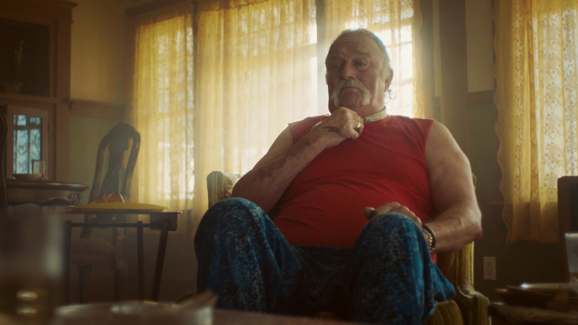 An older man adjusts his collar while sitting in a living room, joined by two younger individuals in a tense domestic setting. The characters engage in a quiet, contemplative moment within a dimly lit home.