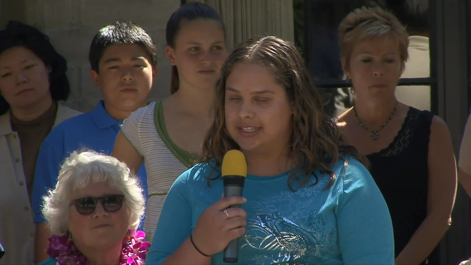 A puppy raiser shares her experience with the audience during a formal ceremony celebrating the graduation of service dogs. She speaks into a microphone while standing before a group of attendees.