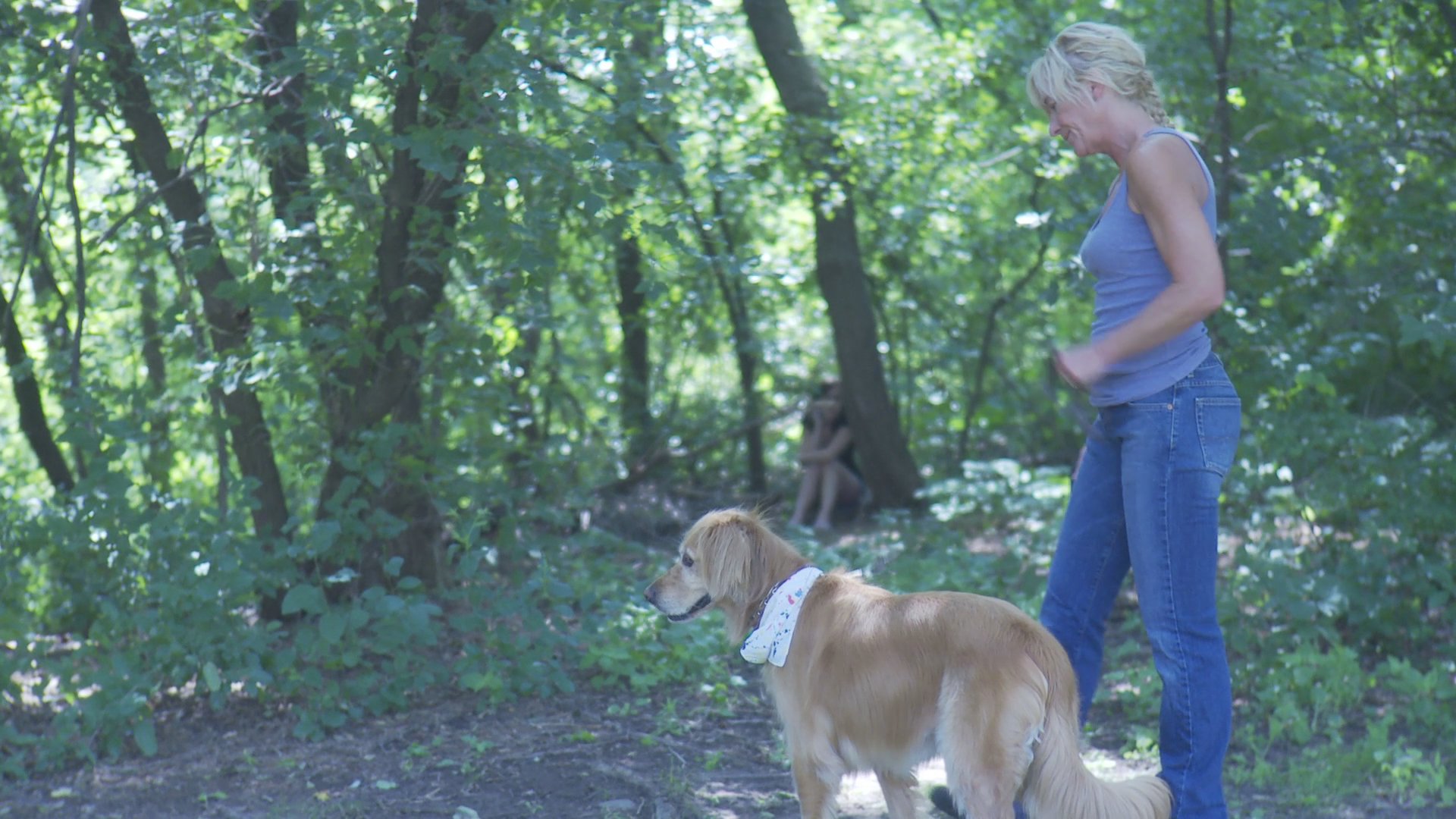 A woman sits against a tree in a wooded area, engaged in an emotional phone conversation. In the background, a person walks a golden retriever through the trees.