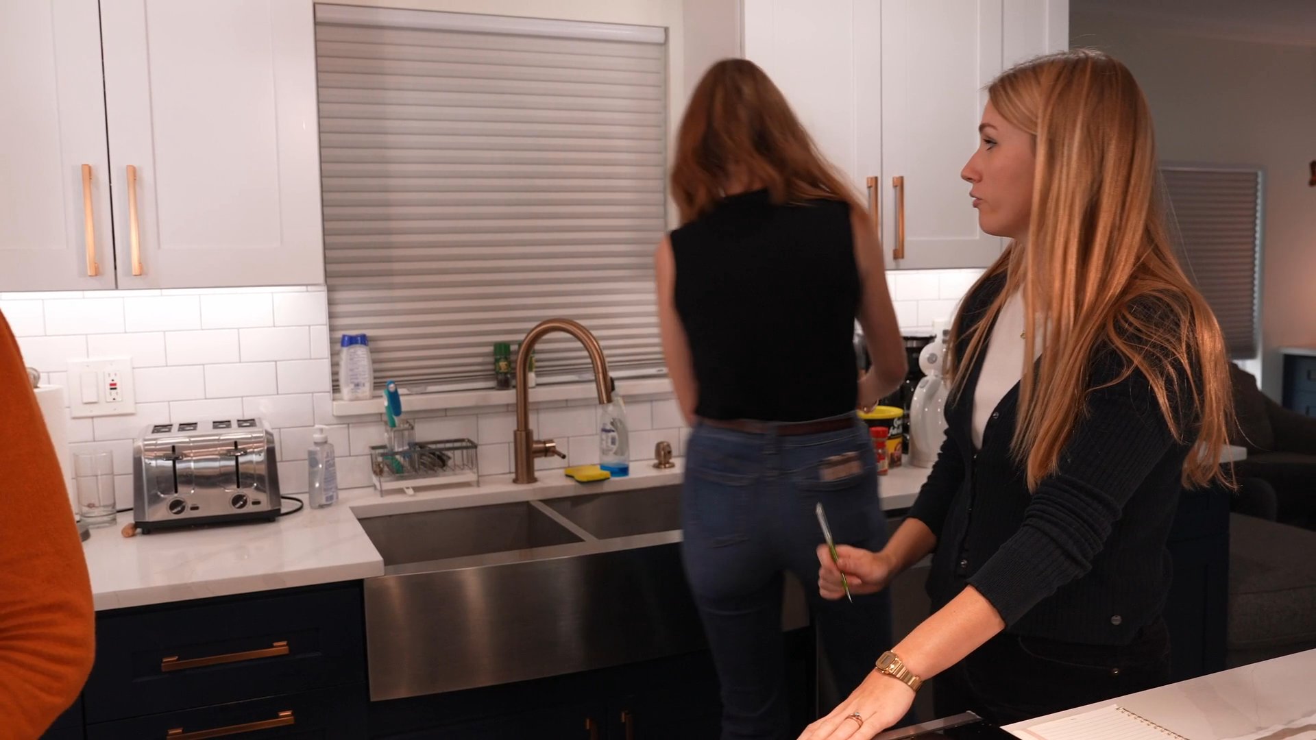 Two women interact while working together in a home kitchen, alternating between tasks and active conversation. The scene focuses on their dynamic as they move about the space.