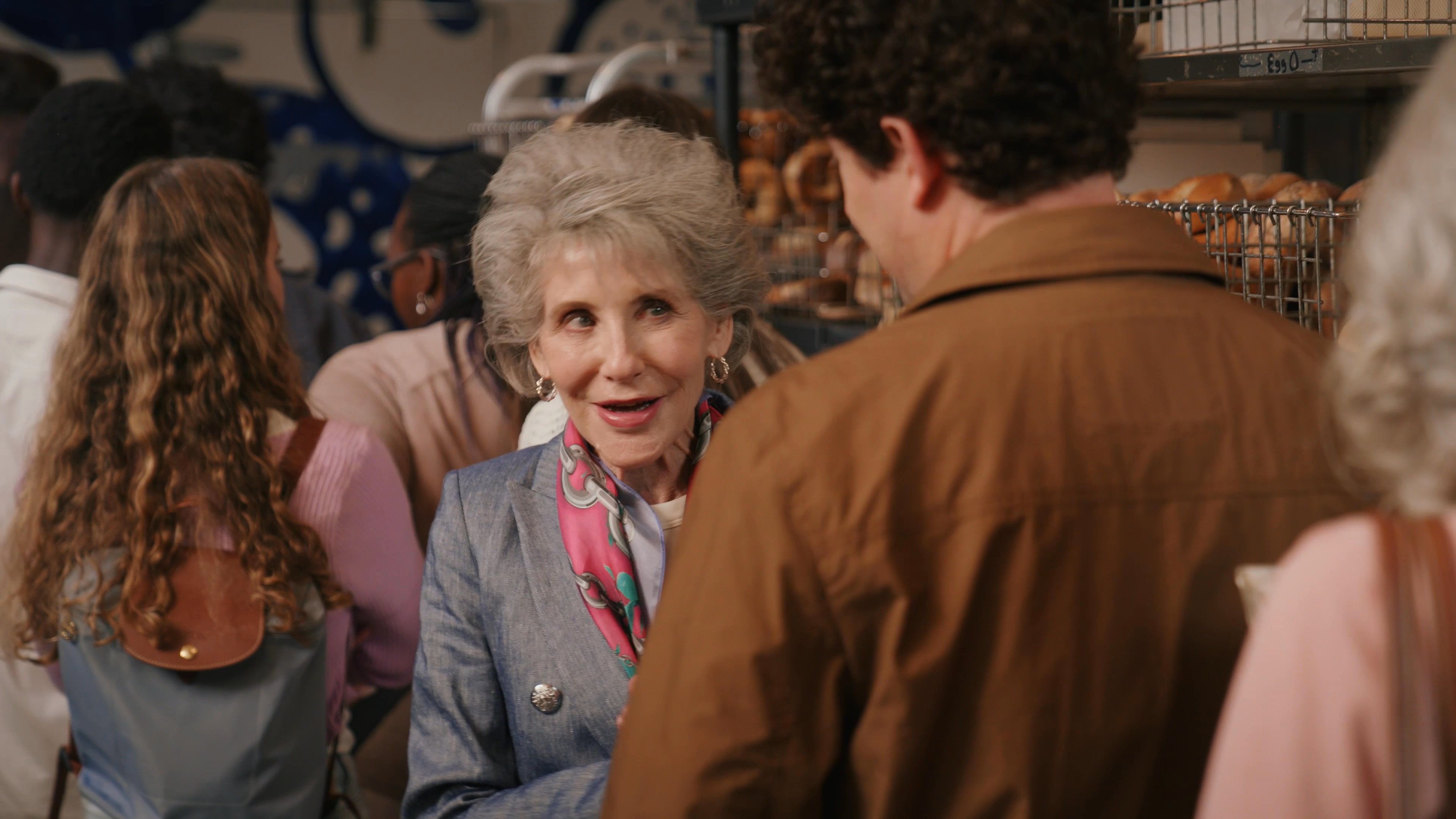 An older woman engages in a pleasant conversation with a bakery employee while standing in front of a rack of bagels.