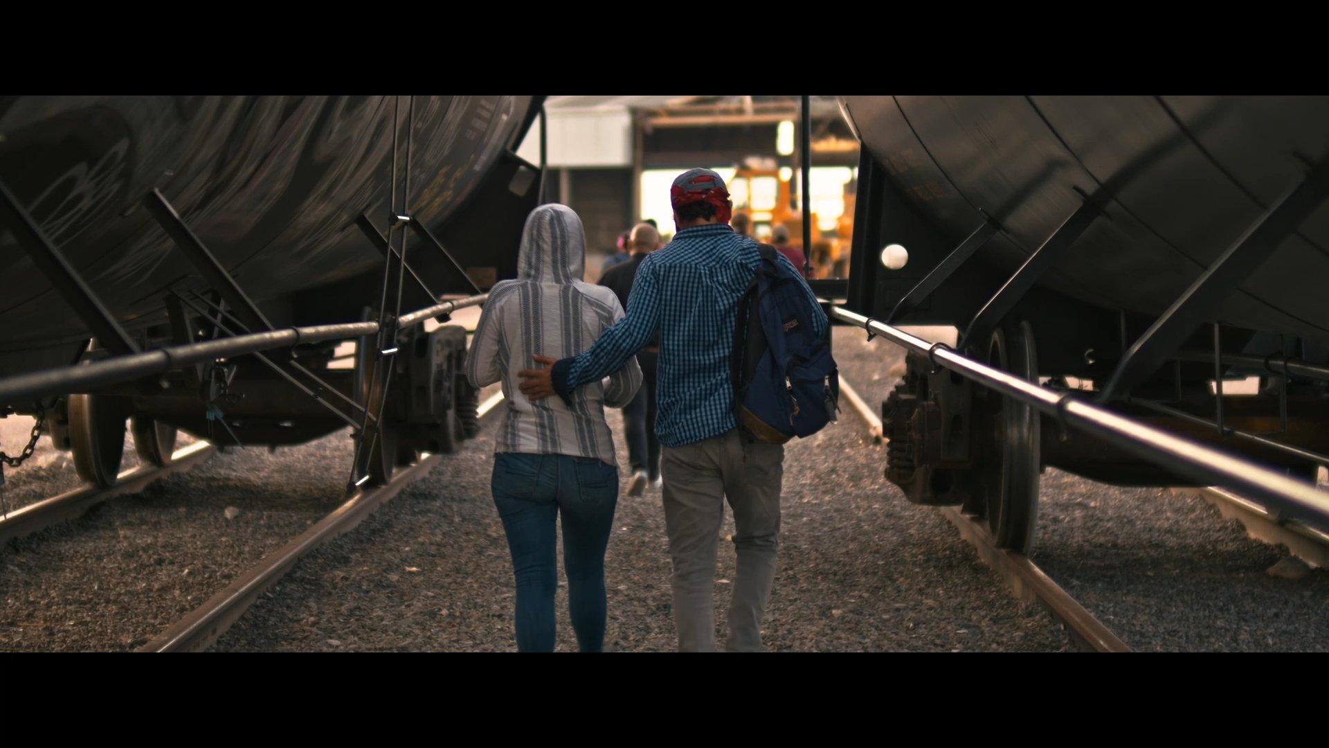 Two groups of individuals navigate a rail yard and a border barrier while discussing the changing landscape of their route. The sequence emphasizes themes of uncertainty and environmental shifts during a migration journey.