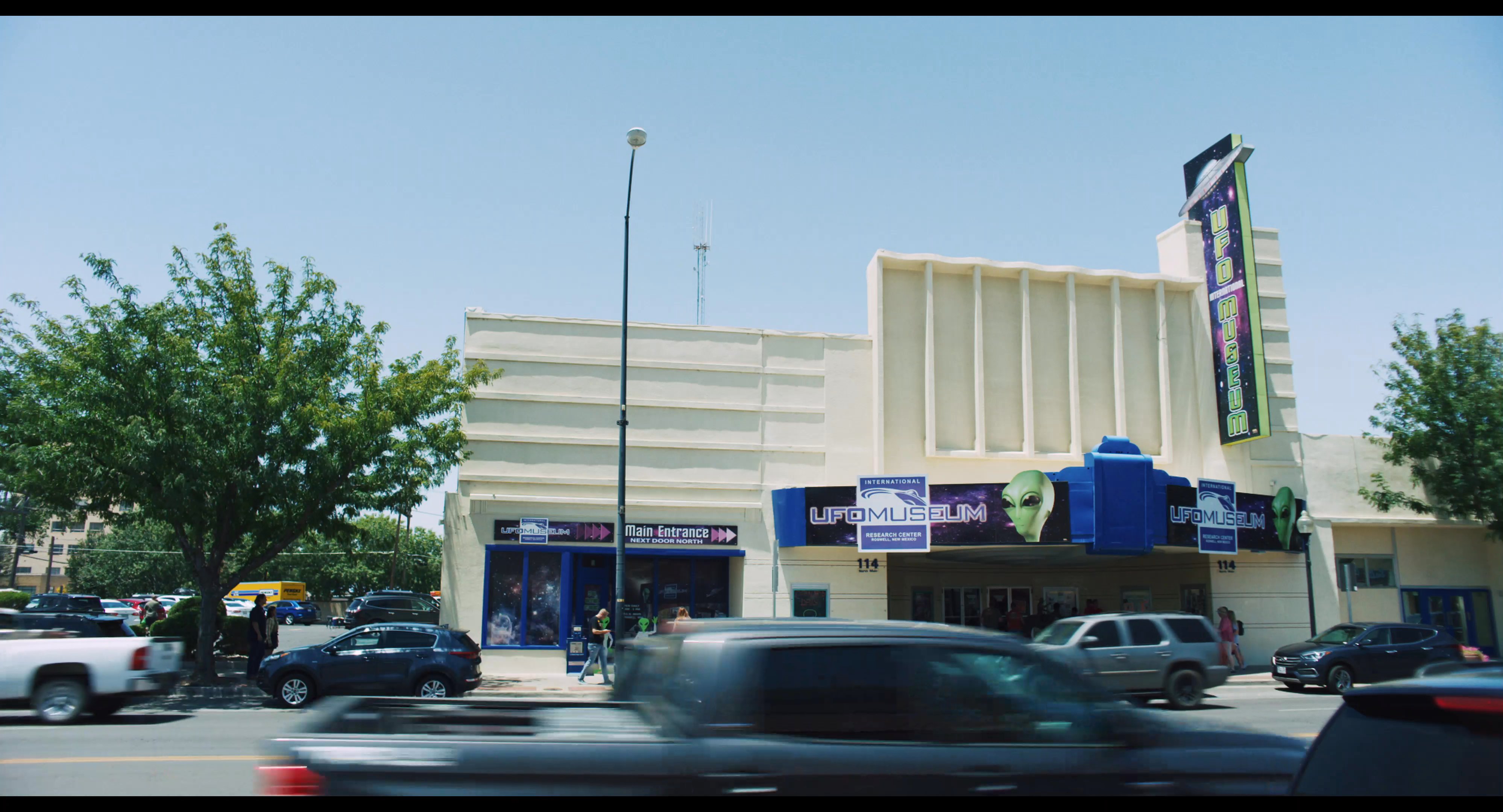 A young man and woman walk together through a sunlit commercial parking area while engaged in conversation. They pass by storefronts and busy street traffic as they observe their surroundings.