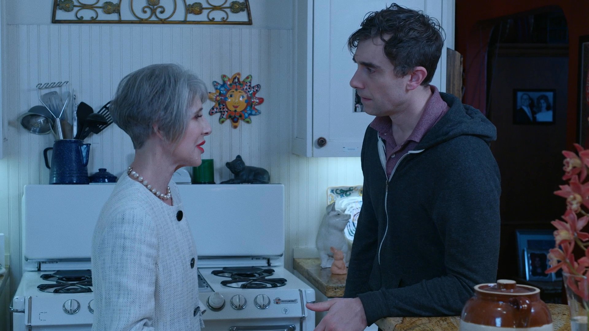 An older woman and a younger man engage in a serious, emotionally charged conversation while standing in a kitchen.