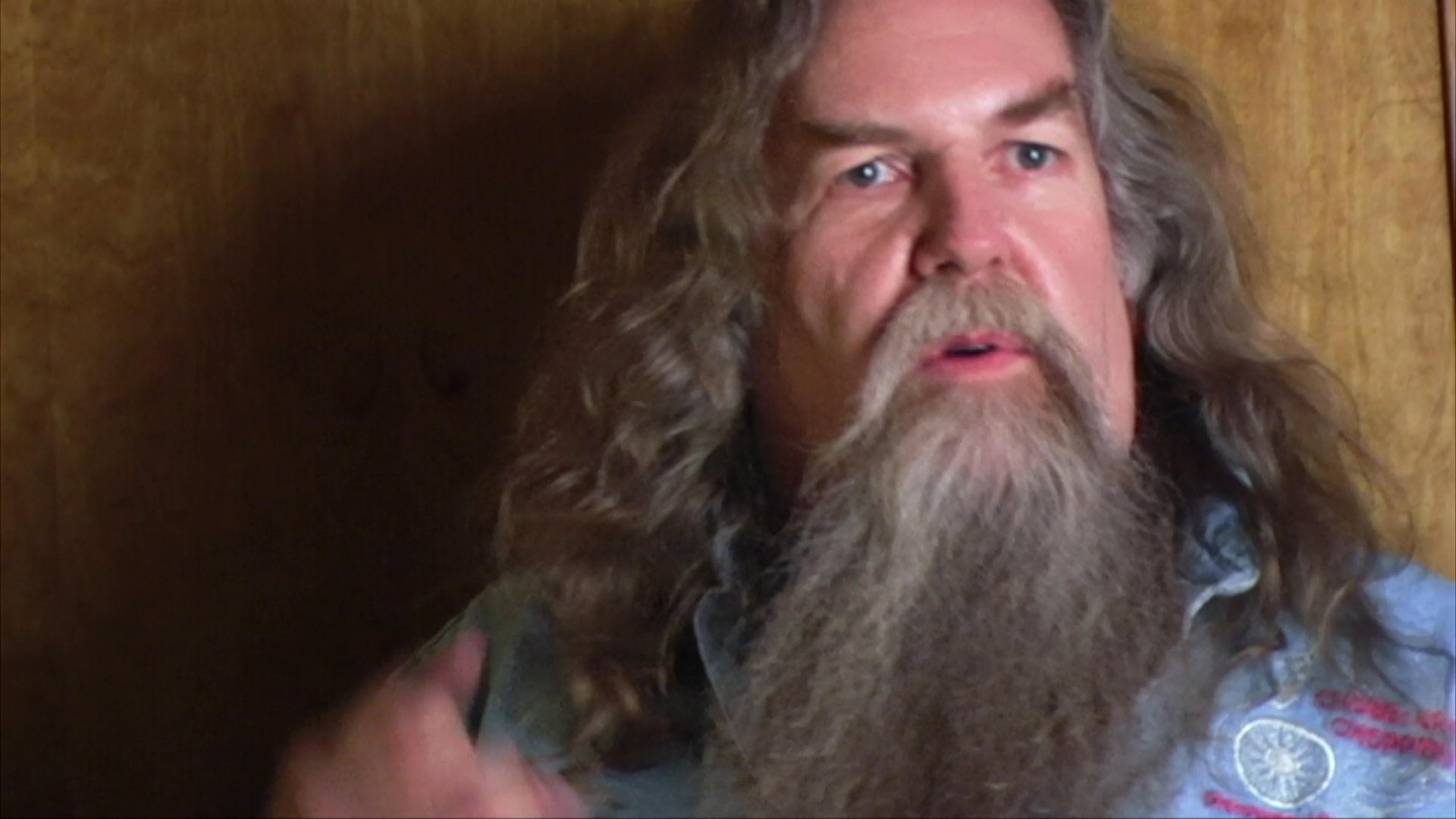 A man with a long beard gestures expressively while speaking directly to the camera against a wood-paneled background.