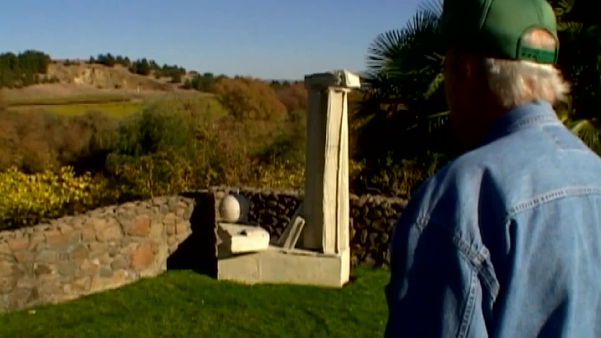 An older man in a green cap walks through a landscaped garden among stone walls, a delicate chair-like sculpture, and a concrete form. He reflects on the appeal of the chair and the concrete piece as a respite from color and human figures.