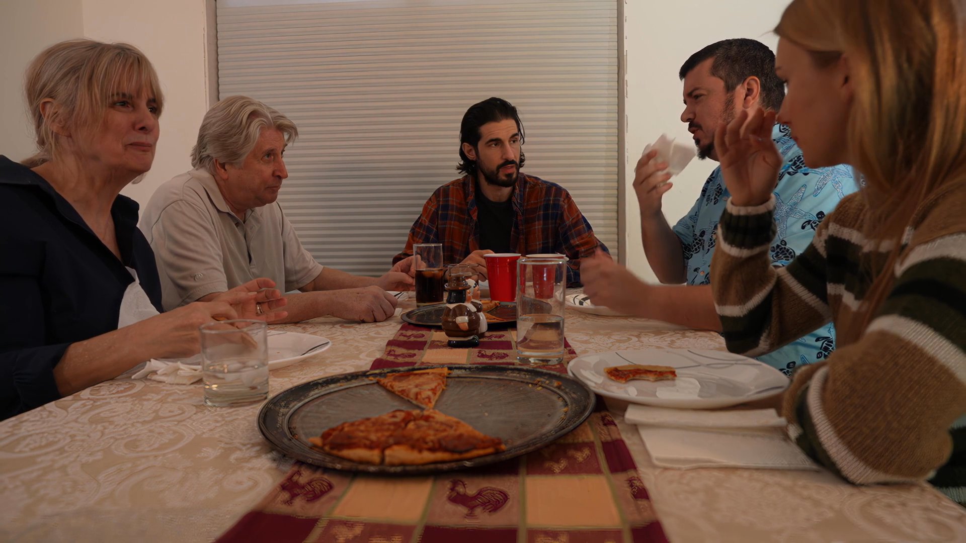 A group of friends enjoys a relaxed evening meal while engaged in conversation around a dining table. The scene transitions to a later moment where a couple rests together in a home setting.