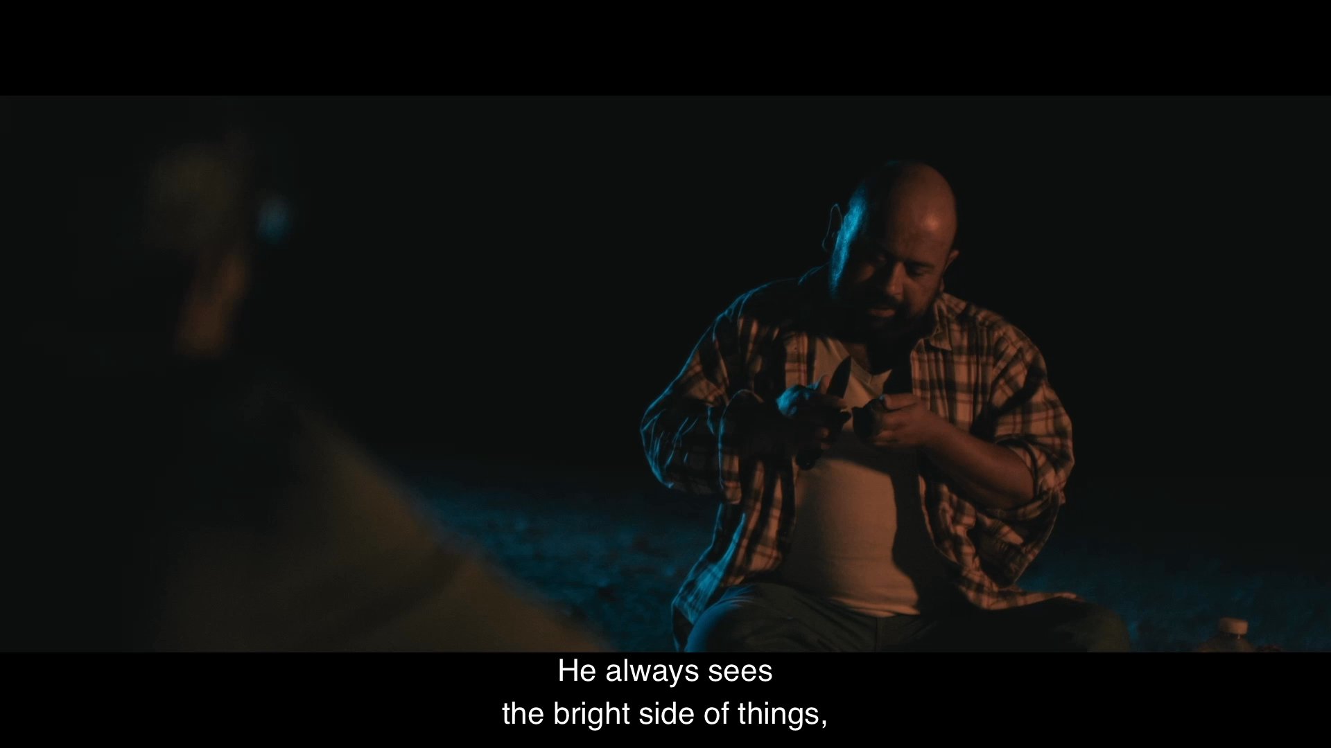 Two men sit together by a campfire at night, sharing personal reflections about family upbringing and the character of a father.