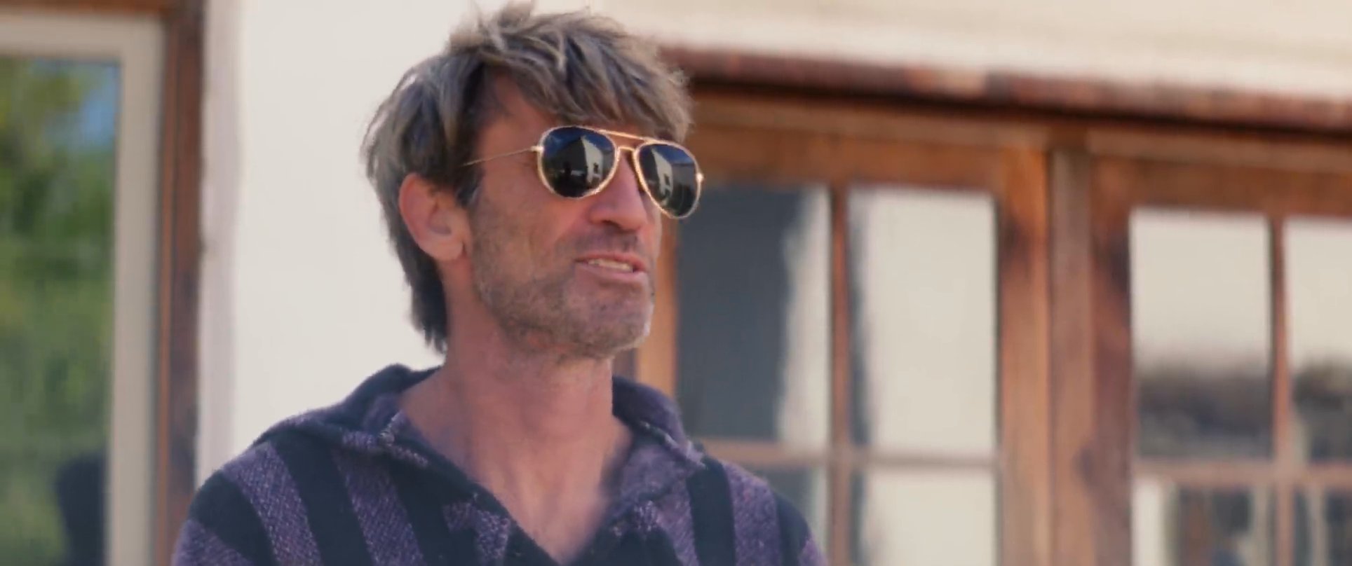 A man in a striped hooded sweater engages in conversation while wearing sunglasses on a patio. He eventually sits down while his interlocutor remains standing.
