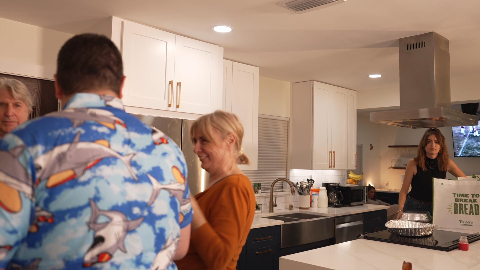 Friends and family socialize and coordinate meal preparations in a modern kitchen decorated for a holiday gathering. The group engages in warm greetings and collaborative tasks during an informal domestic event.