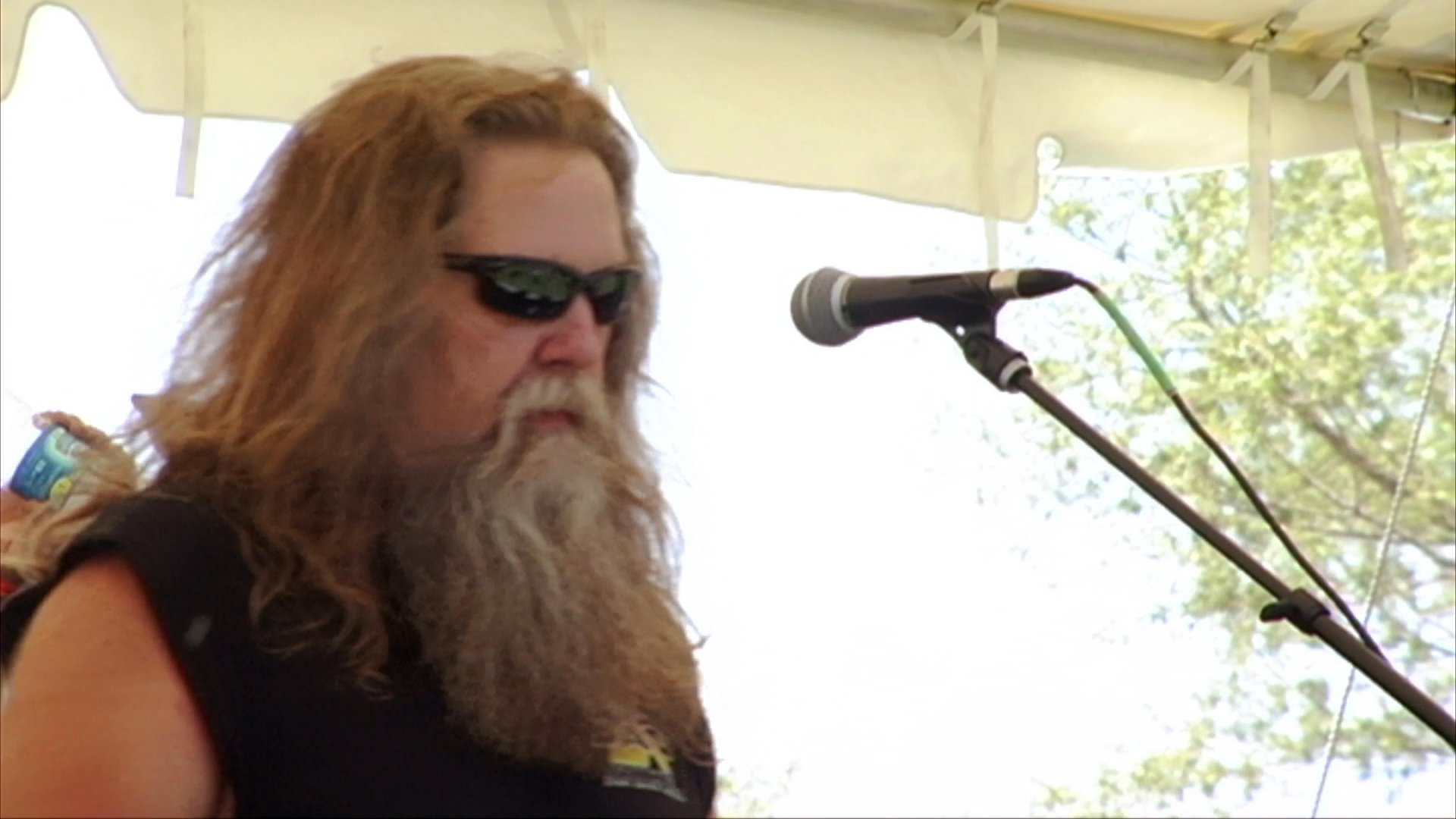 A long-haired, bearded rock musician performs vocals on an outdoor stage during a daytime biker rally event. The band plays instruments while a crowd watches from below the stage canopy.