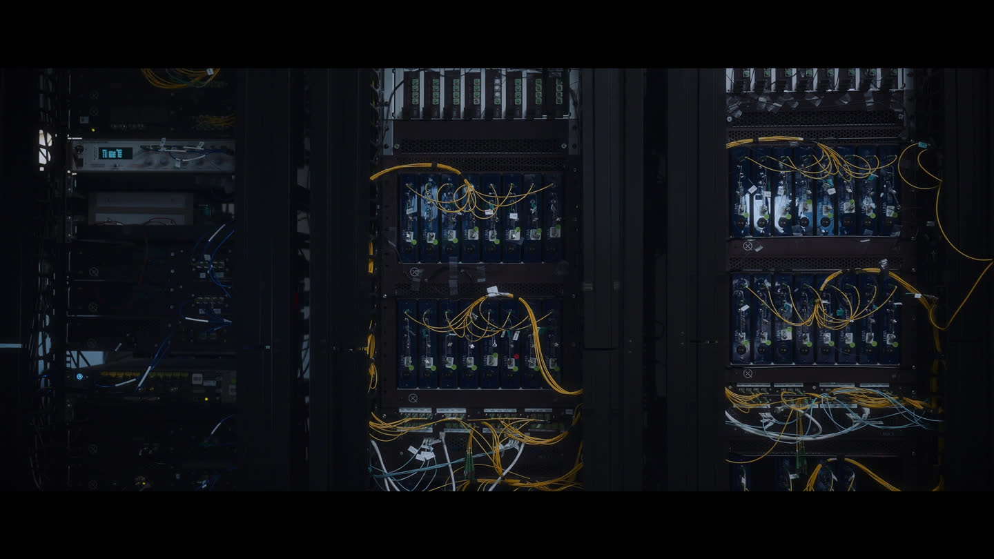 Film still: illuminated equipment racks and connected cables in Xanadu’s laboratory