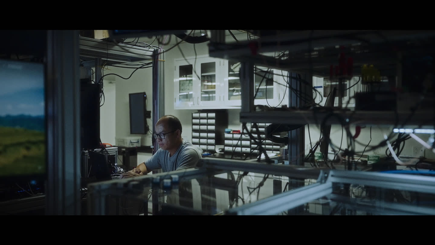 Film still: a team member at a computer surrounded by laboratory equipment