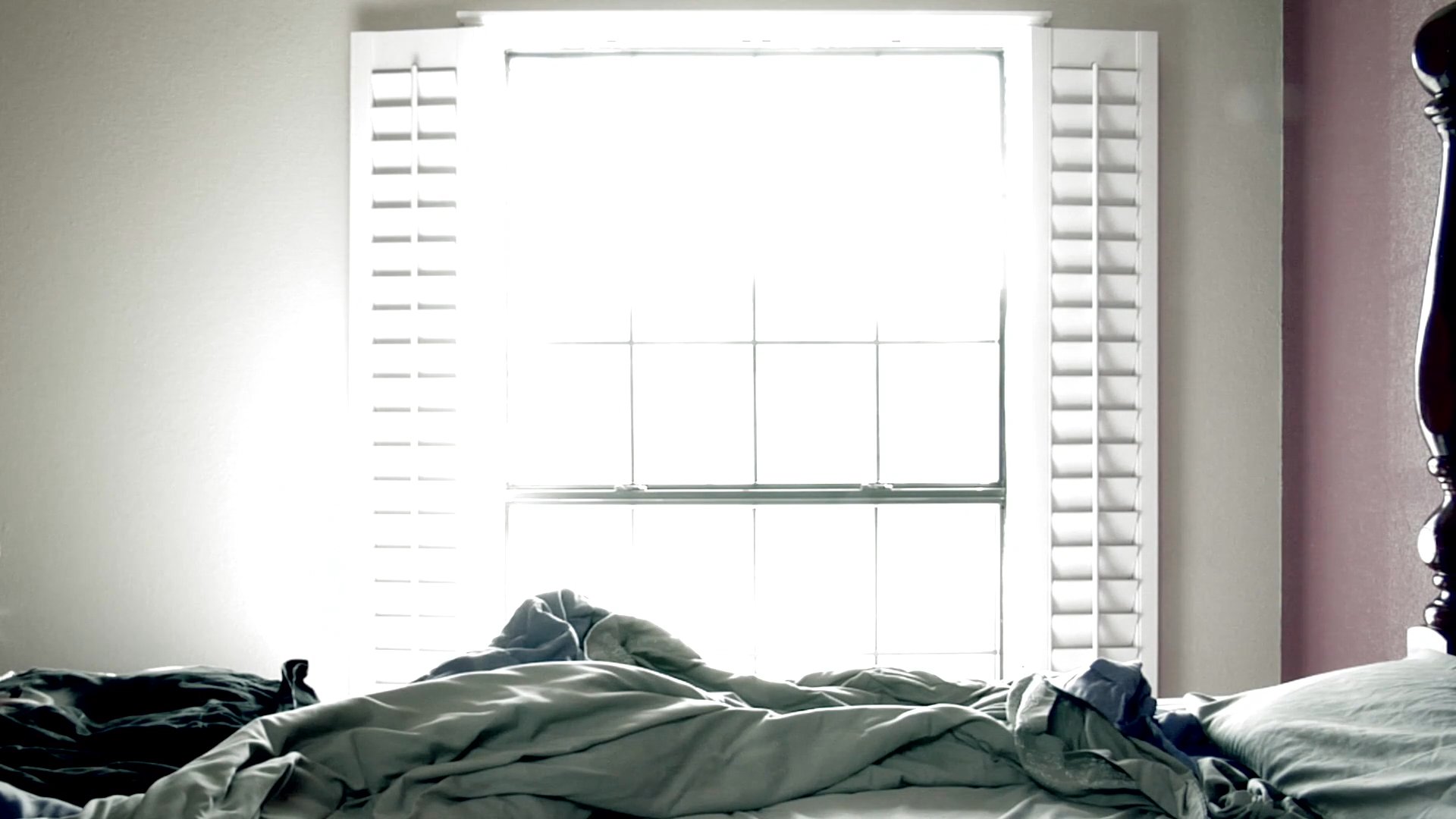 A man in a black shirt stands in a bedroom and looks intently through a window at an outdoor property. The view alternates between his focused expression inside and the sunlit yard outside.