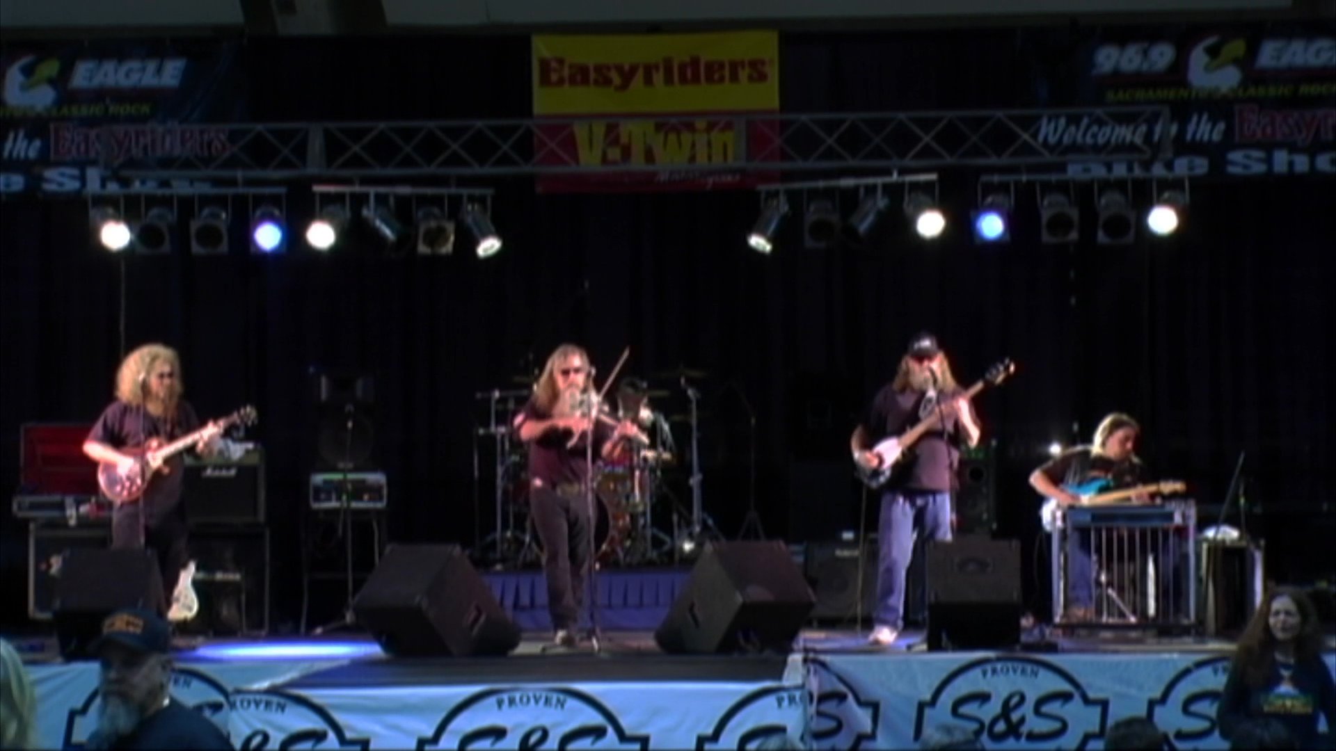 A woman conducts an interview before transitioning to a live band performance on a stage featuring guitar and fiddle instrumentation.