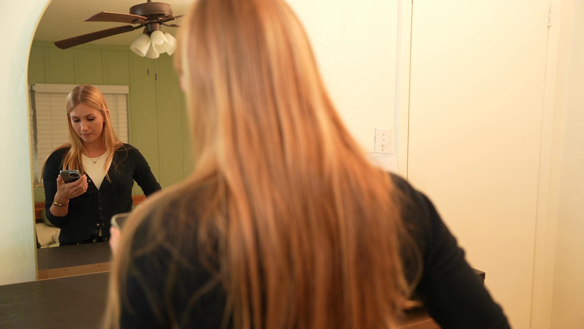 A woman holds a smartphone displaying a video call while interacting with a man in a domestic living space. The scene captures the transition from digital communication to in-person engagement.