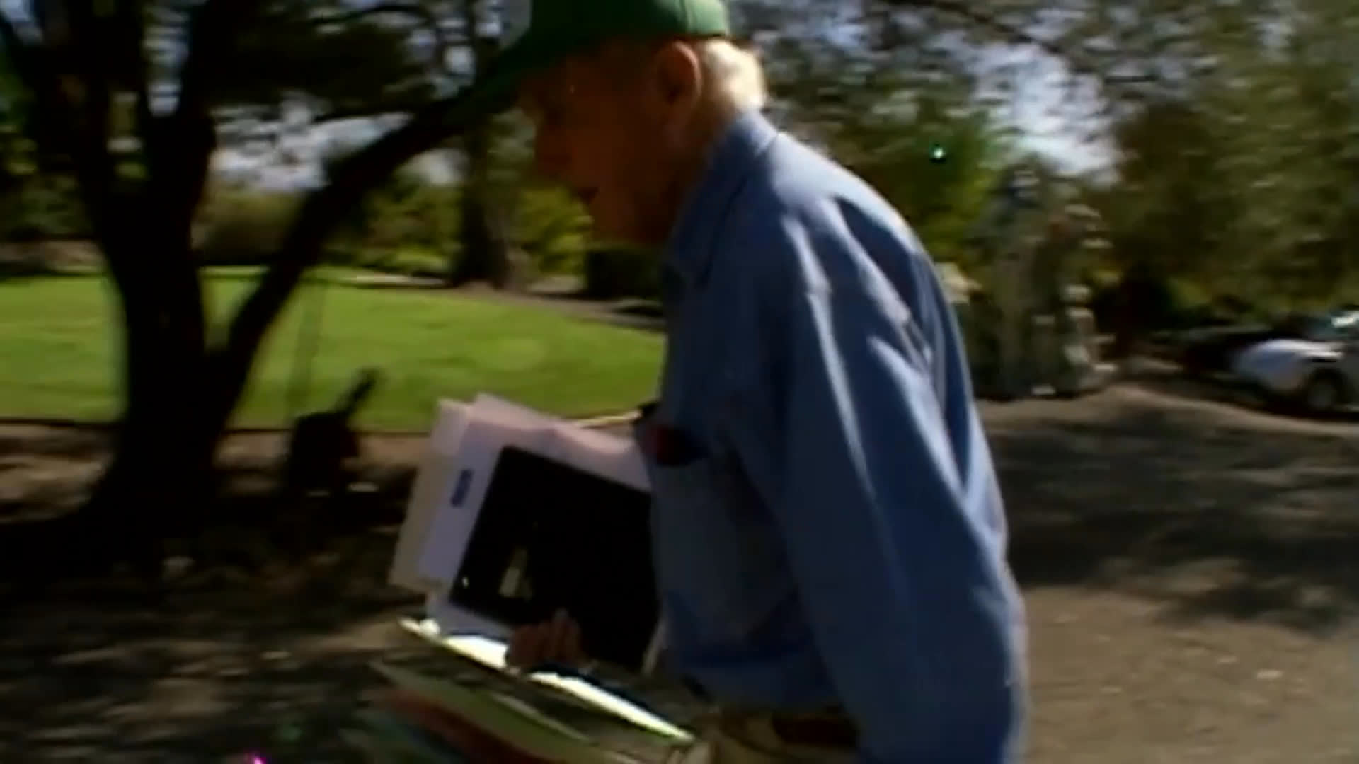 An older man and a woman discuss what they would save from a house fire beside a large pile of assorted objects. He then carries a rectangular item across leafy grounds and enters a furnished room.
