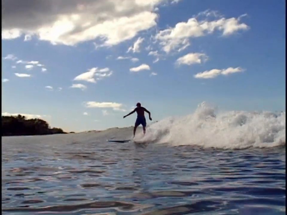 A man practices surfing on open ocean waves, transitioning to a domestic scene where he and his family look through a vintage photo album together. The sequence juxtaposes an active lifestyle with moments of intimate intergenerational reflection.
