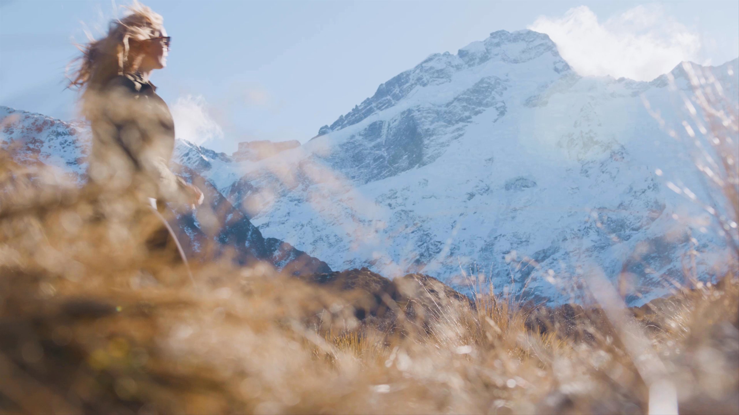 Video of man and woman hiking through a grassy mountain landscape with snowy mountains in the background