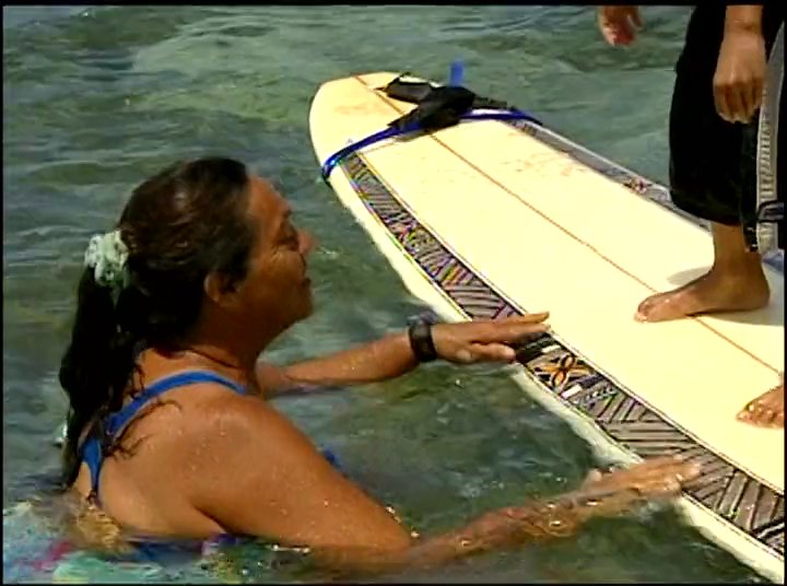 A surf instructor provides guidance and physical support to a young boy as he practices balancing on a surfboard in shallow coastal waters. The session transitions from technique training near the shoreline to the boy riding small waves.