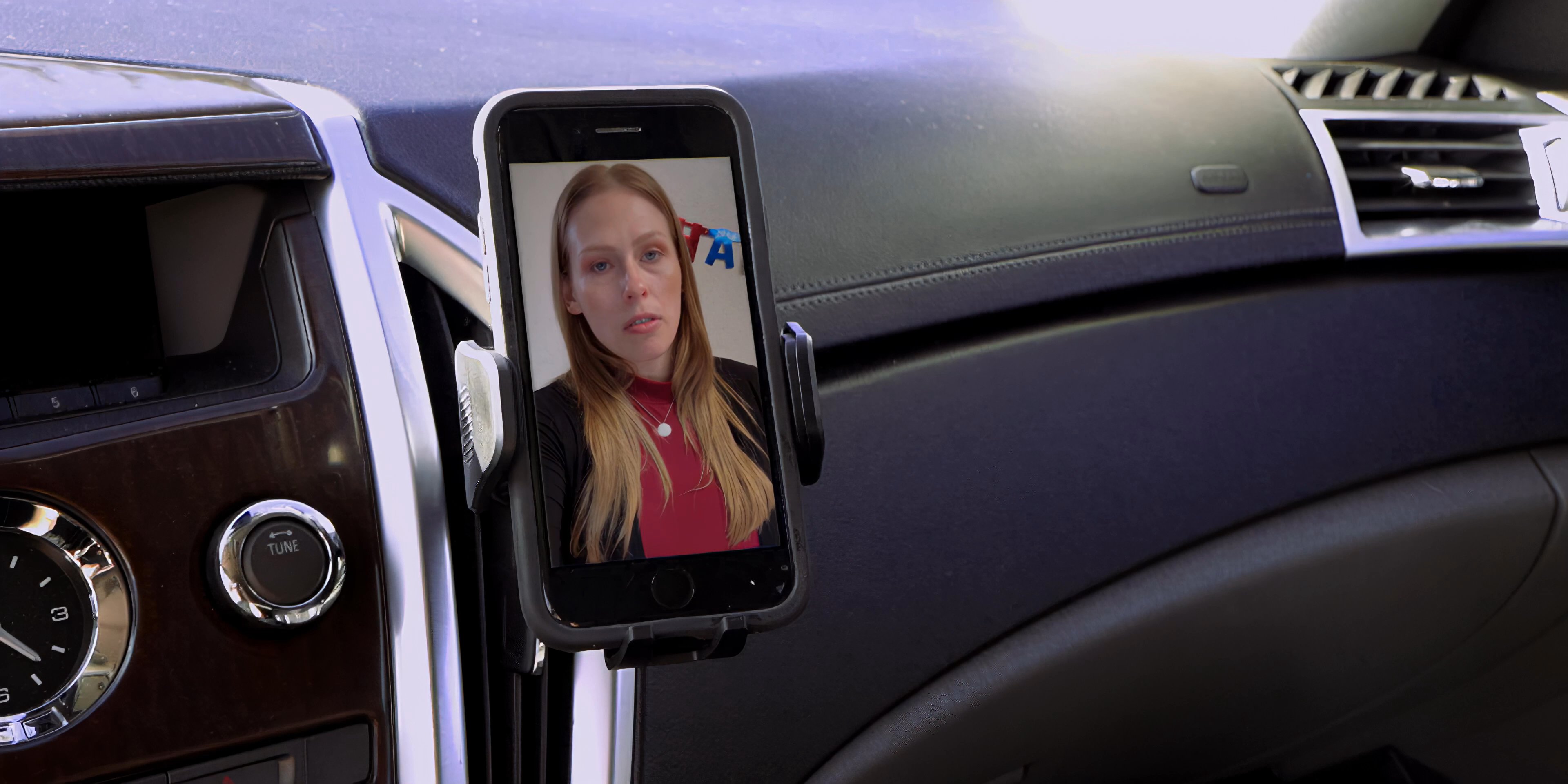 A driver engages in a video call on a smartphone mounted to his car dashboard while parked near a tropical beach. The screen displays alternating participants during the ongoing conversation.