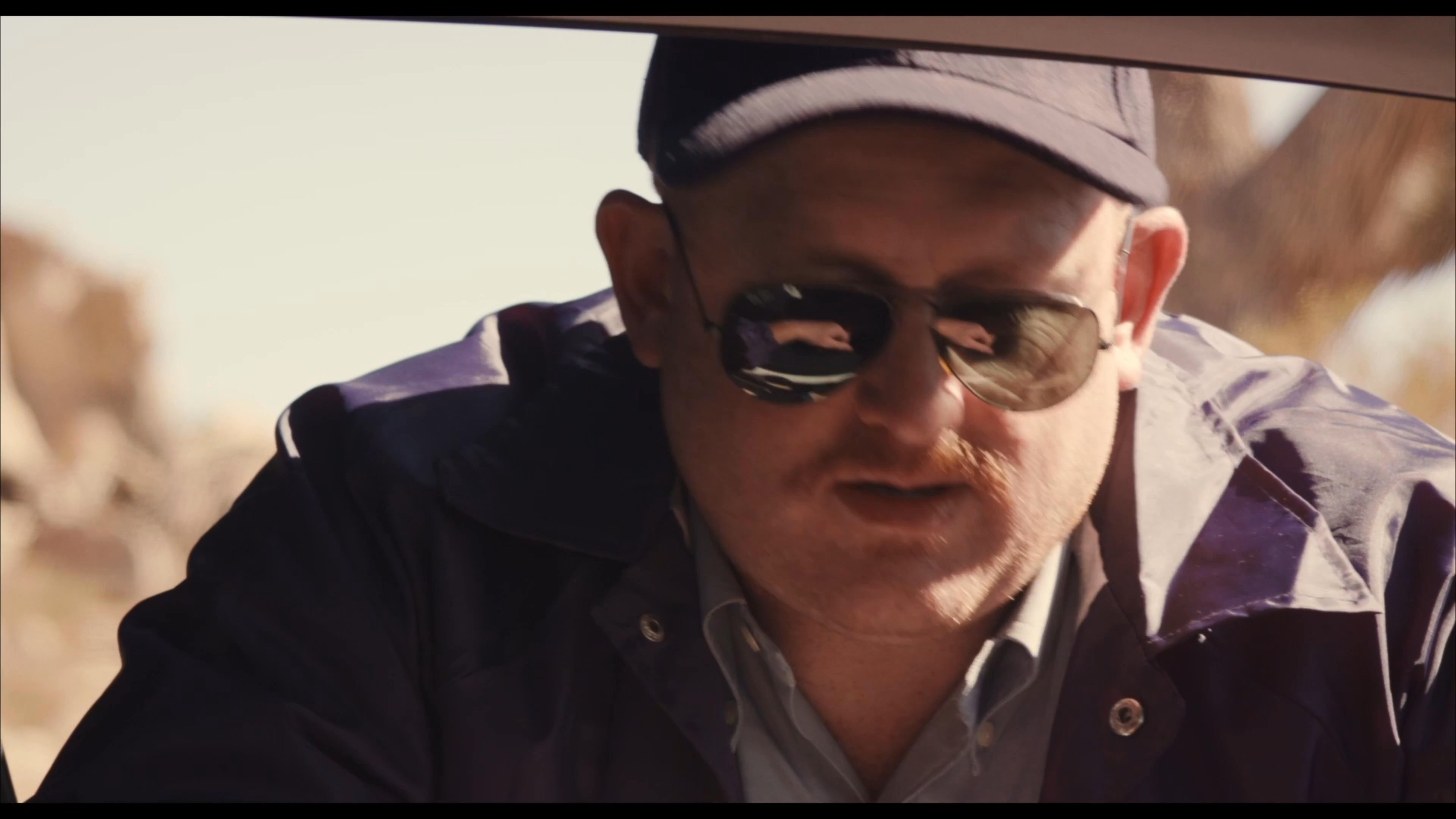 A law enforcement officer conducts a traffic stop with two individuals in a desert setting. The driver and officer engage in a tense interaction during the roadside inspection.