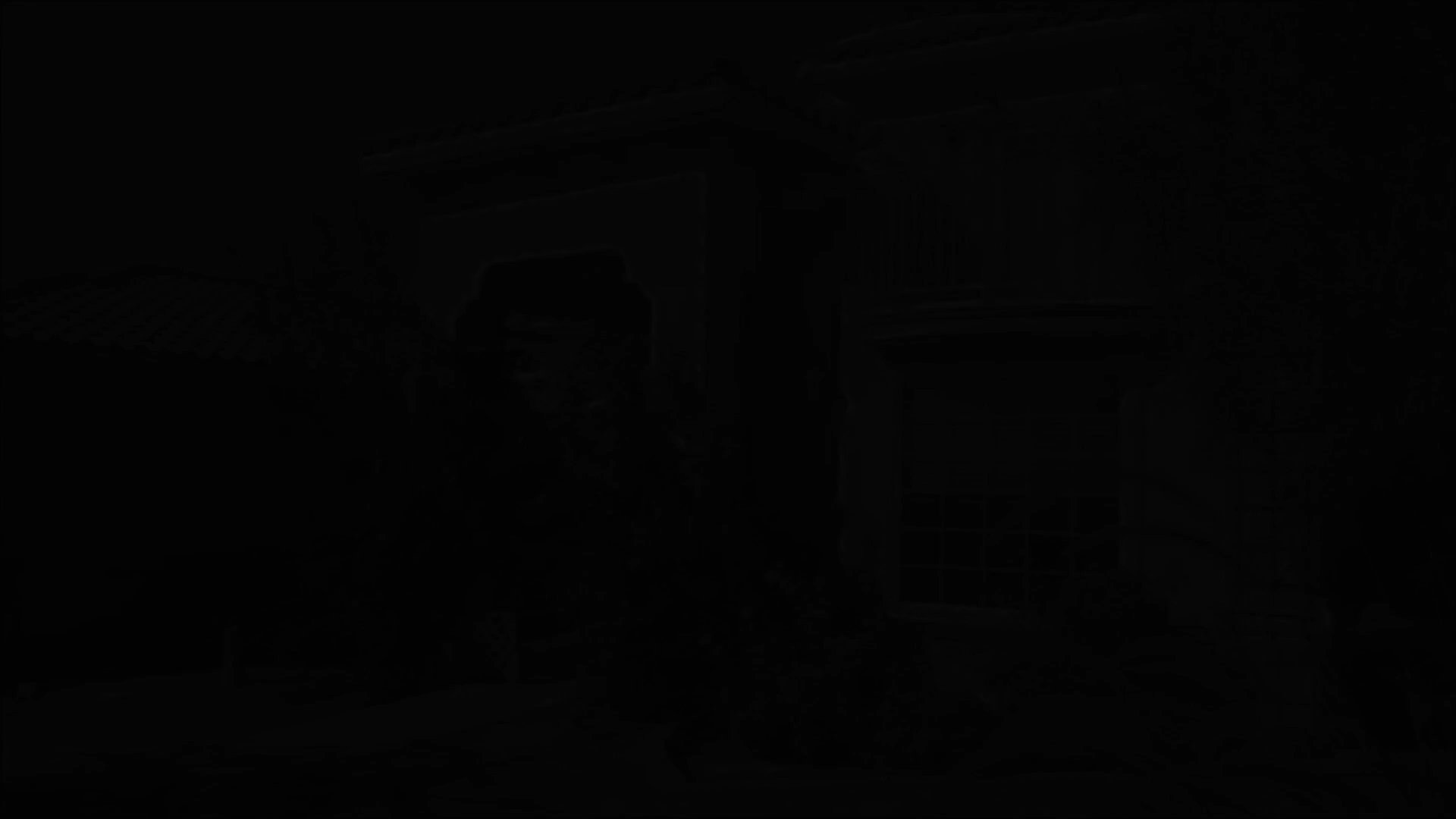 An operative lying in tall grass conducts surveillance on a residential property before two individuals approach the perimeter wall to continue their tactical mission.