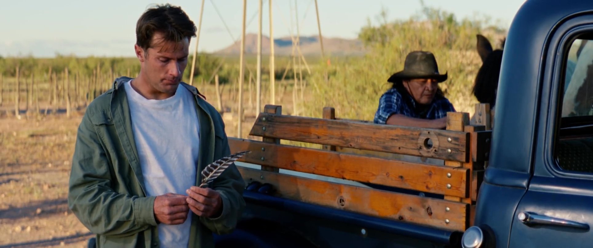 An Indigenous elder offers guidance to a young man, presenting him with a ceremonial feather near a vintage pickup truck on a horse ranch.