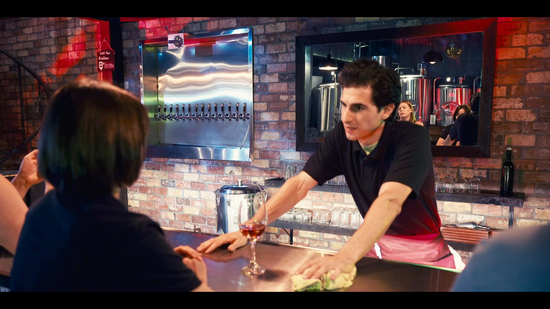 A bartender interacts with a customer while efficiently cleaning and organizing the service counter. He maintains a professional demeanor within the industrial setting of a brewery or taproom.