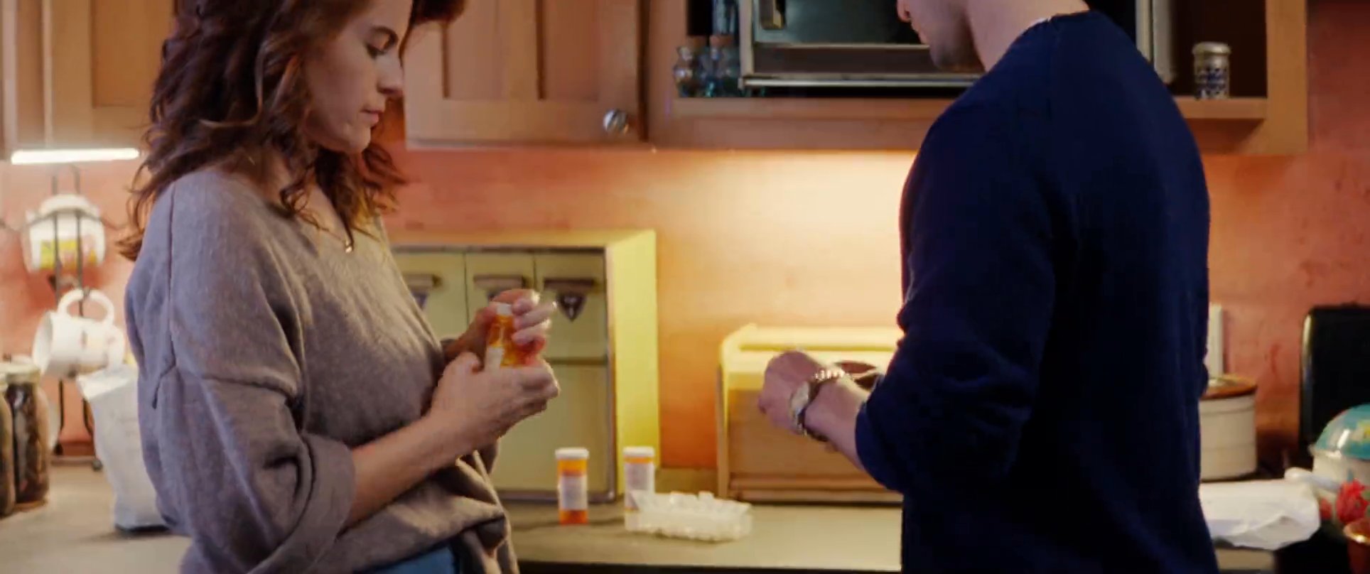 A man in sunglasses receives a book outdoors before shifting to a domestic setting where a man and woman consult over a medicine bottle in a kitchen. The scene highlights a moment of personal care and quiet communication within a home.